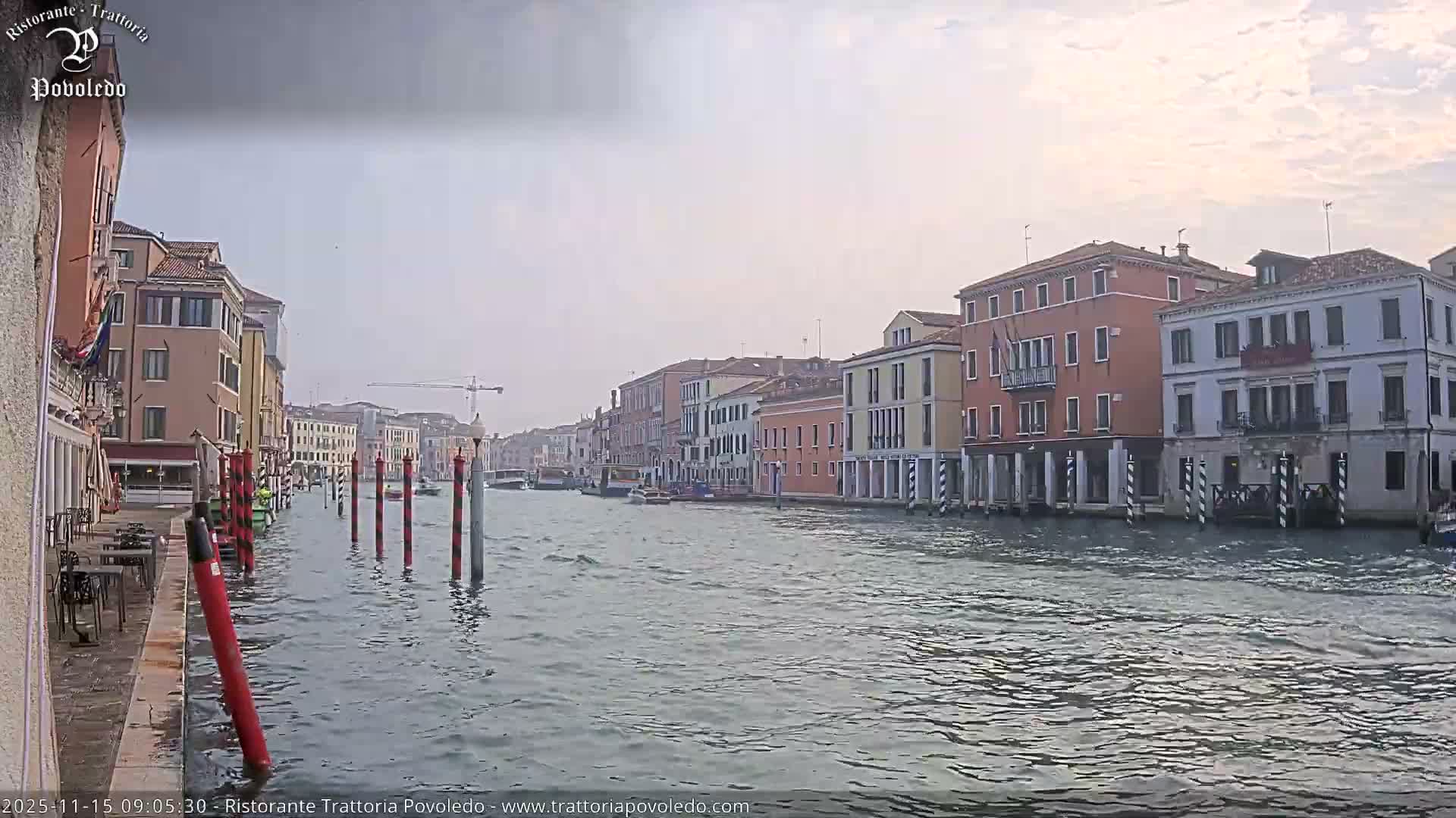 A wide canal in Venice, lined with colorful buildings and a waterside cafe on the left, features several boats and mooring poles on the water under a cloudy sky.