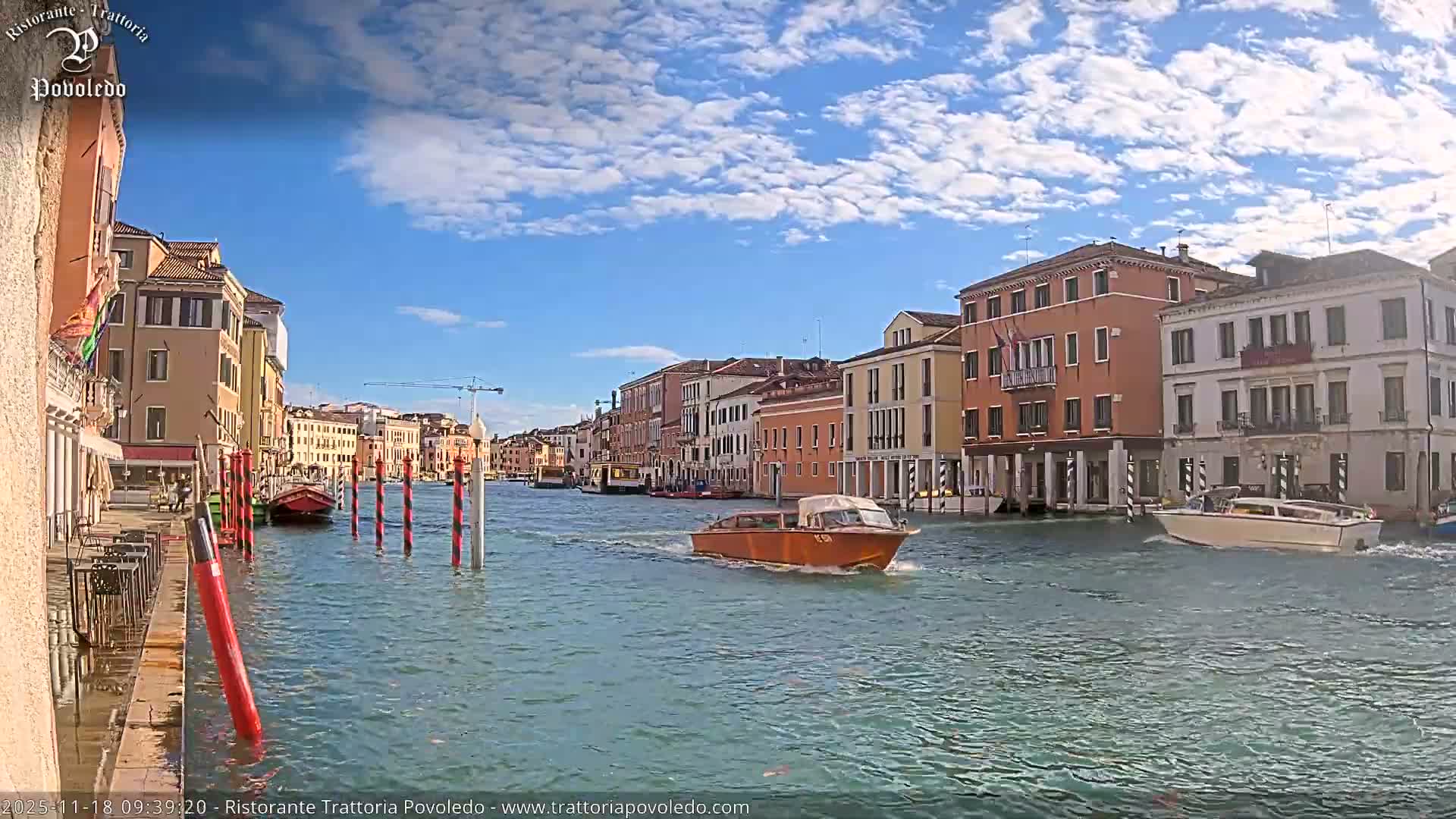 An orange motorboat cruises along a sunlit canal lined with colorful historic buildings under a clear blue sky dotted with fluffy white clouds on a bright day in Venice.