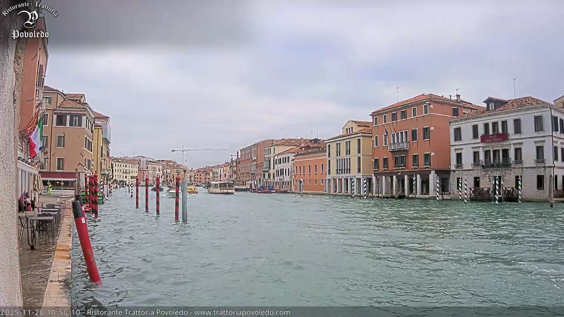 A bustling canal in Venice is lined with colorful historic buildings under an overcast sky, as boats navigate the rippling water where the high tide partially submerges the waterside path.
