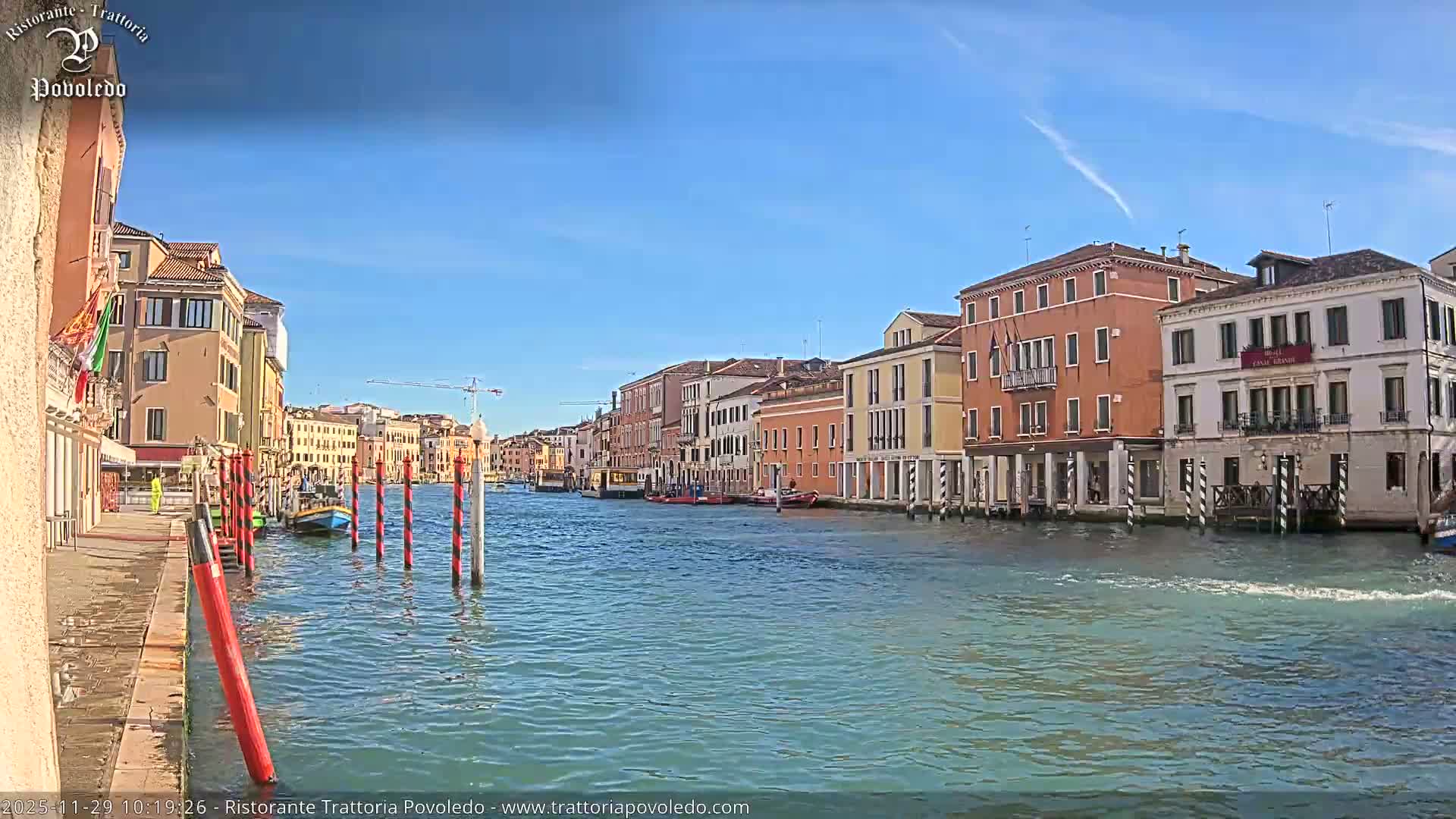 Under a clear, sunny blue sky, a wide canal is flanked by a continuous row of colorful, multi-story historic buildings and dotted with boats and striped mooring poles.