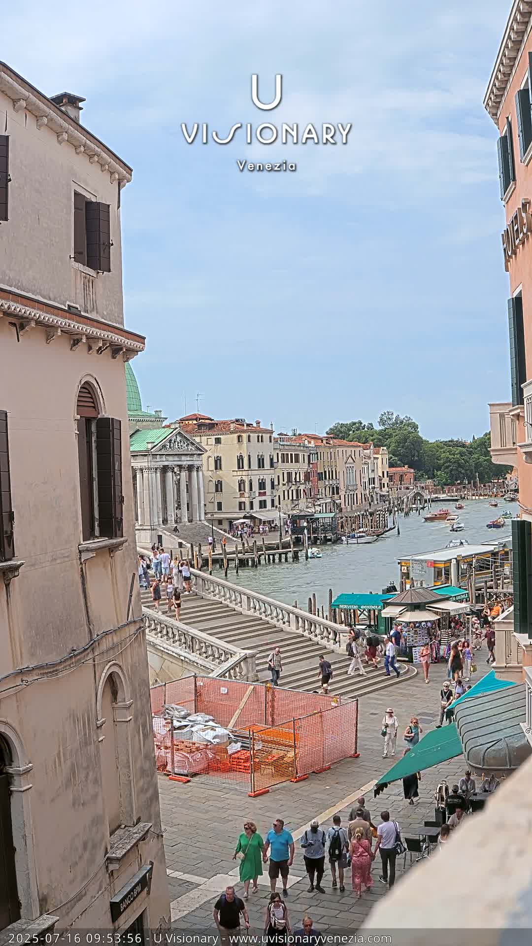 A sunny day in Venice shows a canal with boats, buildings lining the waterway, and many people walking along a stone staircase and the waterfront.