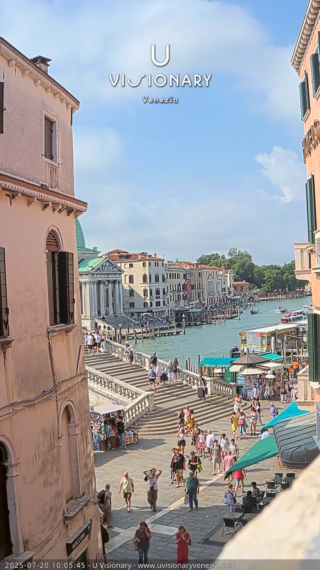 A sunny day in Venice shows a canal with boats, buildings lining the waterway, and many people walking on a stairway leading down to the water's edge.