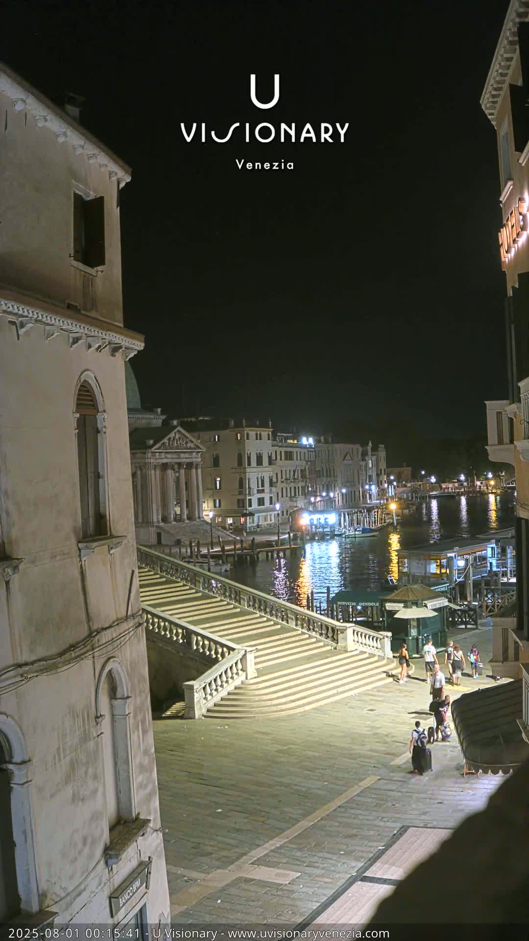 A nighttime view of a canal in Venice, with a long staircase leading down to the water, where people are walking along the waterfront and boats are moored.