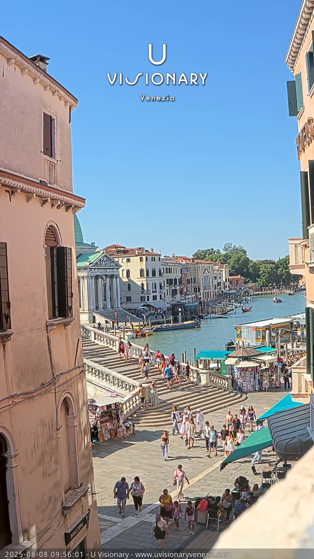 A sunny day in Venice shows a wide view of a canal with boats and buildings, along with many people walking up and down a large staircase leading to a waterfront area with shops and cafes.