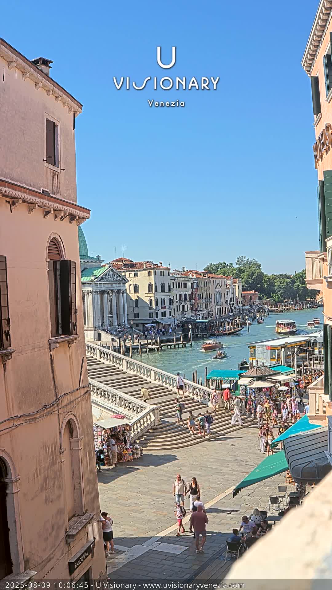 A sunny day in Venice shows a wide shot of a canal with boats, buildings, a stone staircase leading down to the water, and people walking along the waterfront.