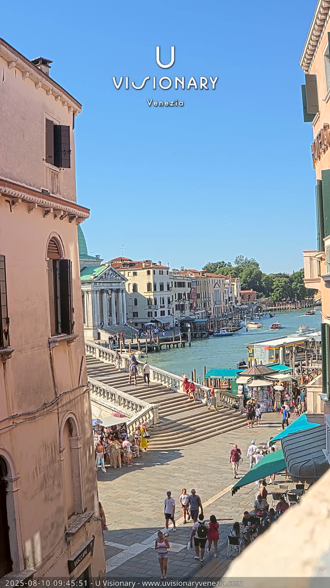 A sunny view of a canal in Venice, showing a stairway leading down to a waterfront area with pedestrians and shops, boats on the water, and buildings lining the canal.