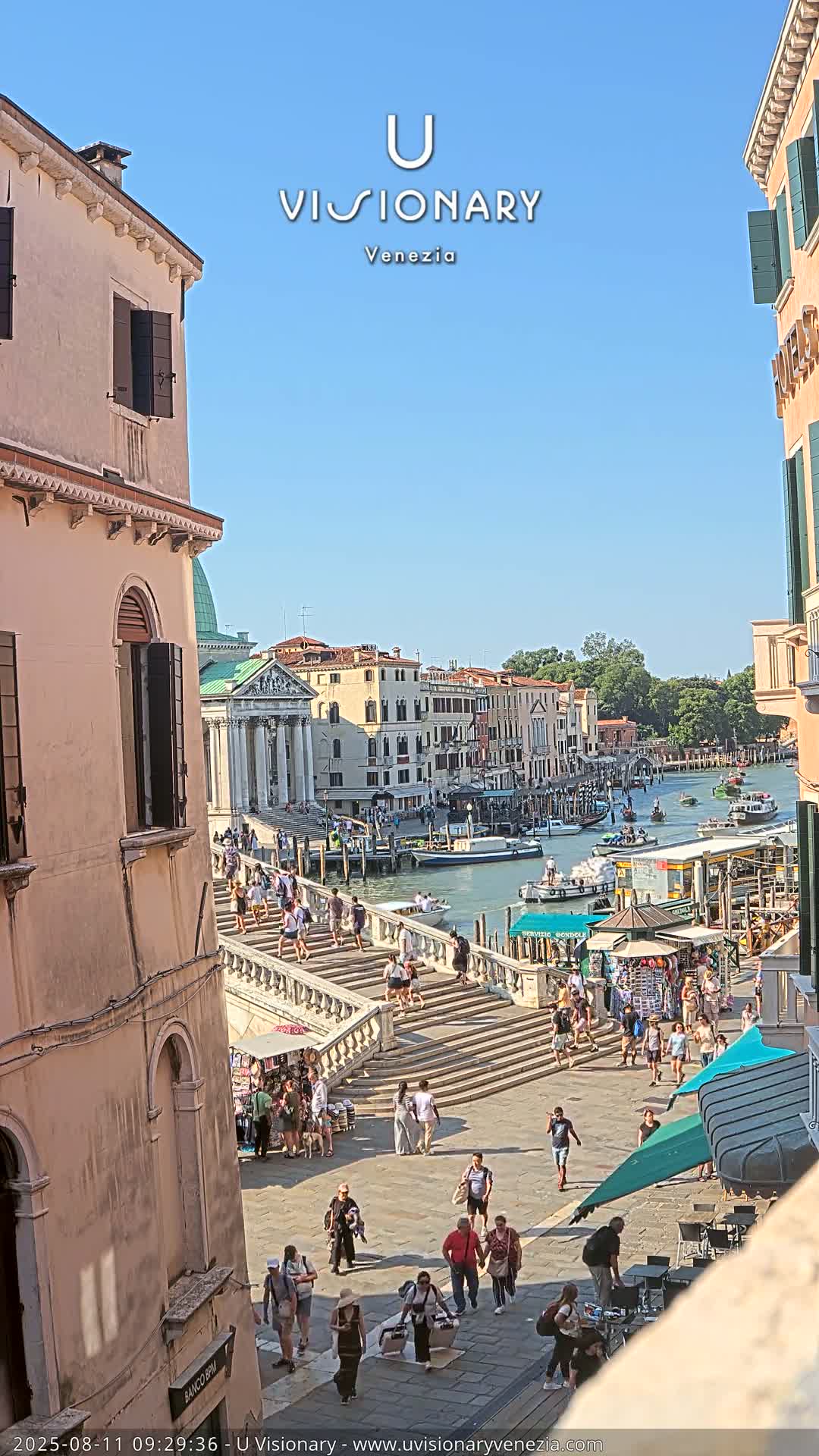 A sunny day in Venice shows a wide staircase descending to a canal, crowded with people and boats, with buildings lining the waterway.