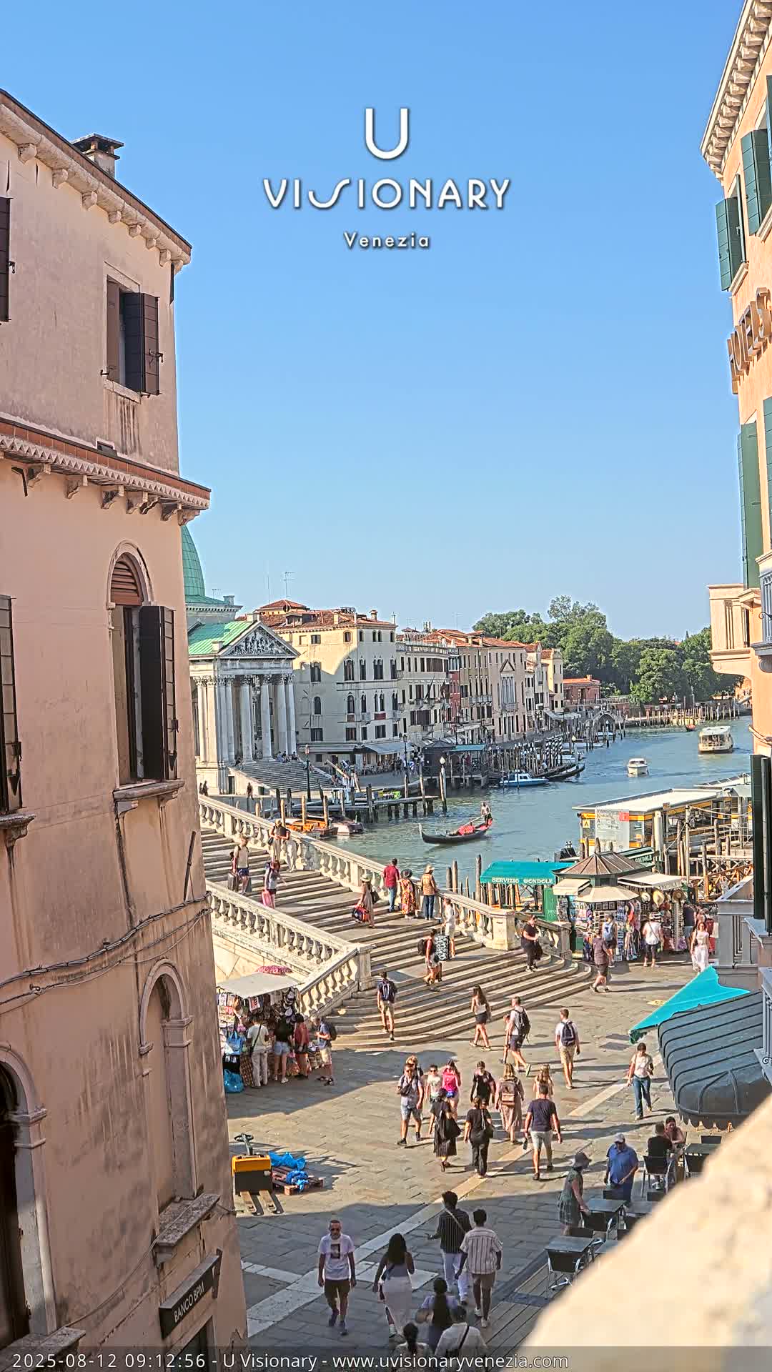 A sunny day in Venice shows a wide staircase descending towards a canal, crowded with pedestrians and boats, with buildings lining both sides of the waterway.