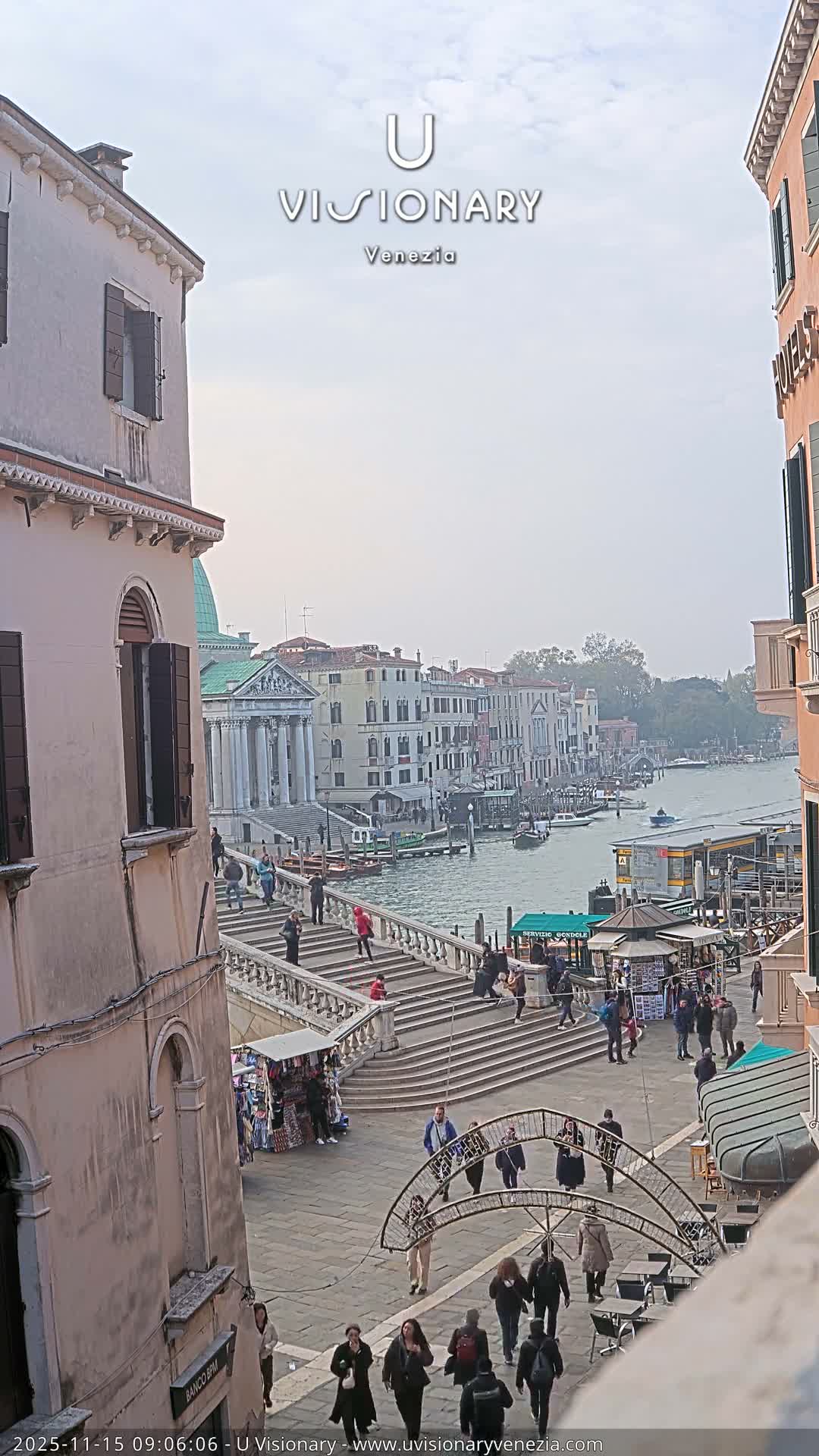 On a hazy, overcast day, a wide Venetian canal is bordered by historic buildings and a stone bridge bustling with people, with boats traversing the water and a lively plaza in the foreground.