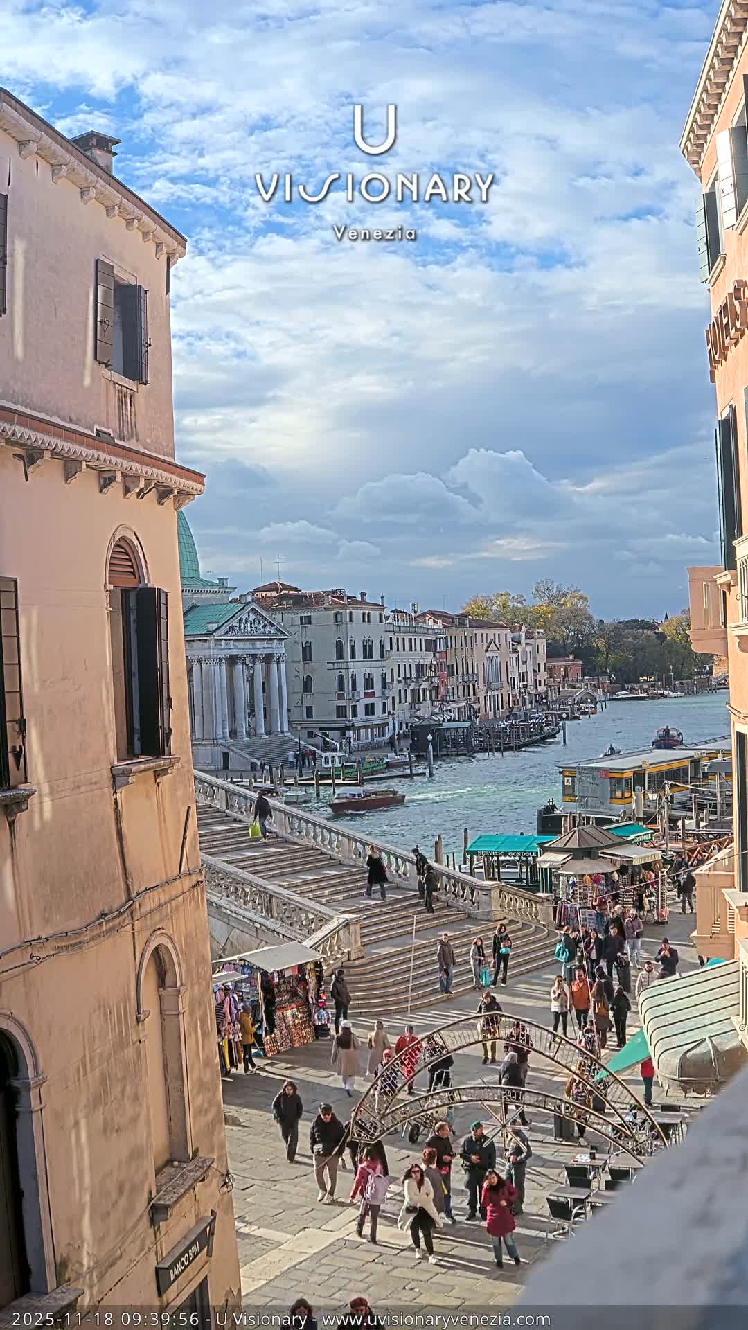 A vibrant outdoor scene in Venice unfolds under a partly cloudy sky, featuring a grand stone staircase teeming with pedestrians descending to a canal bustling with boats and surrounded by classic European architecture.