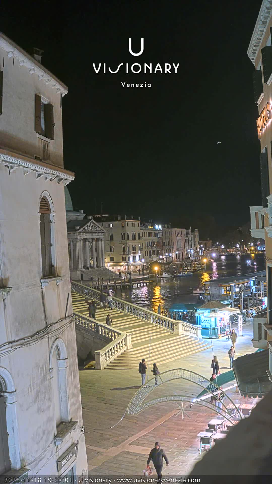 A vibrant outdoor scene in Venice unfolds under a partly cloudy sky, featuring a grand stone staircase teeming with pedestrians descending to a canal bustling with boats and surrounded by classic European architecture.