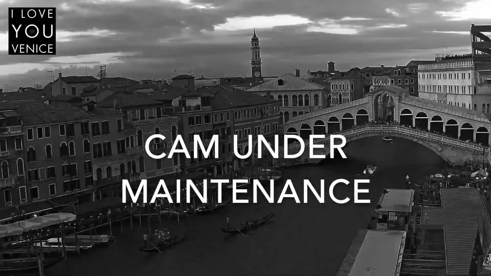 A black and white, cloudy daytime scene depicts a busy Venetian canal with gondolas, historic buildings, and an ornate arched bridge teeming with pedestrians.