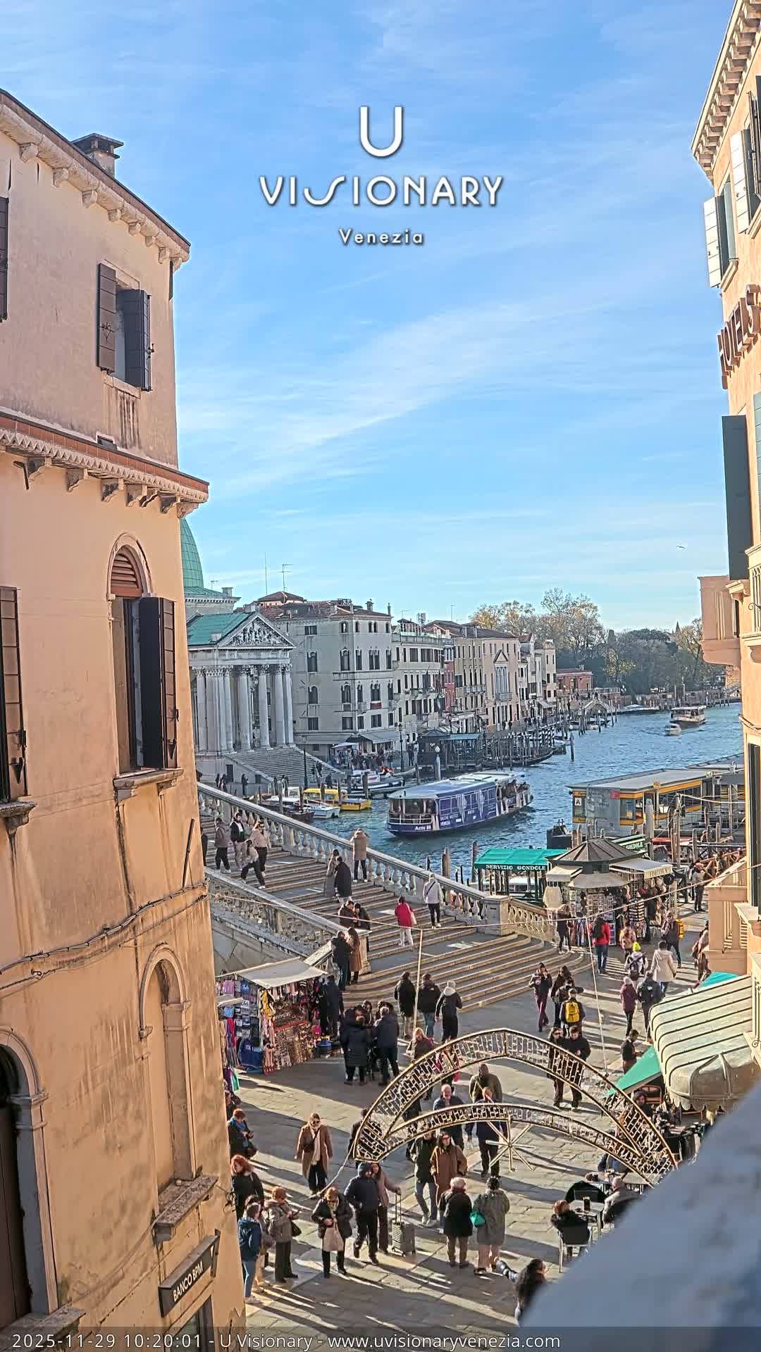 Under a clear blue sky, a sunny daytime view of Venice captures a bustling Grand Canal with boats and historic buildings, alongside a stone bridge crowded with people, market stalls, and festive decorations.