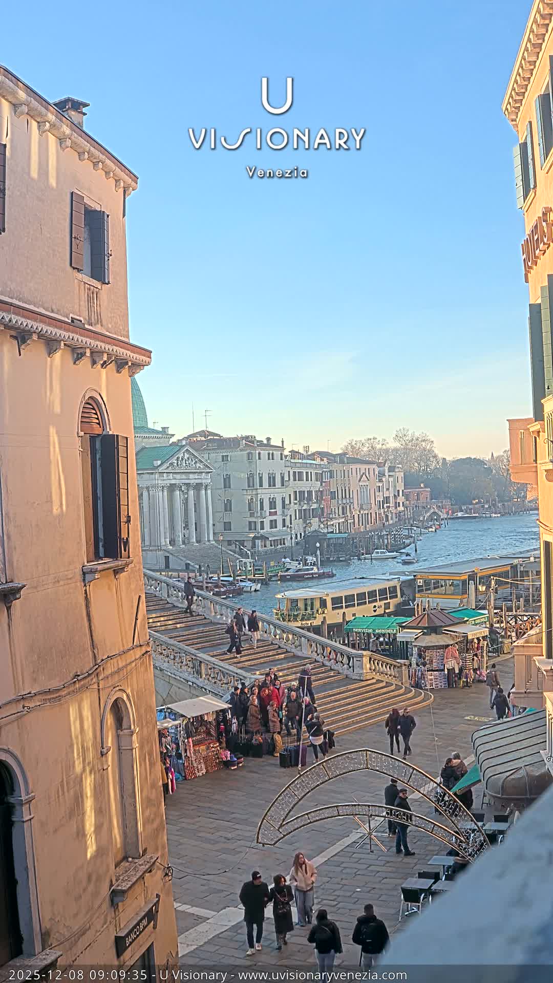 On a clear and sunny morning, a bustling scene in Venice captures people ascending and descending a grand stone bridge over a canal filled with boats, while market stalls and pedestrians animate the paved area below.