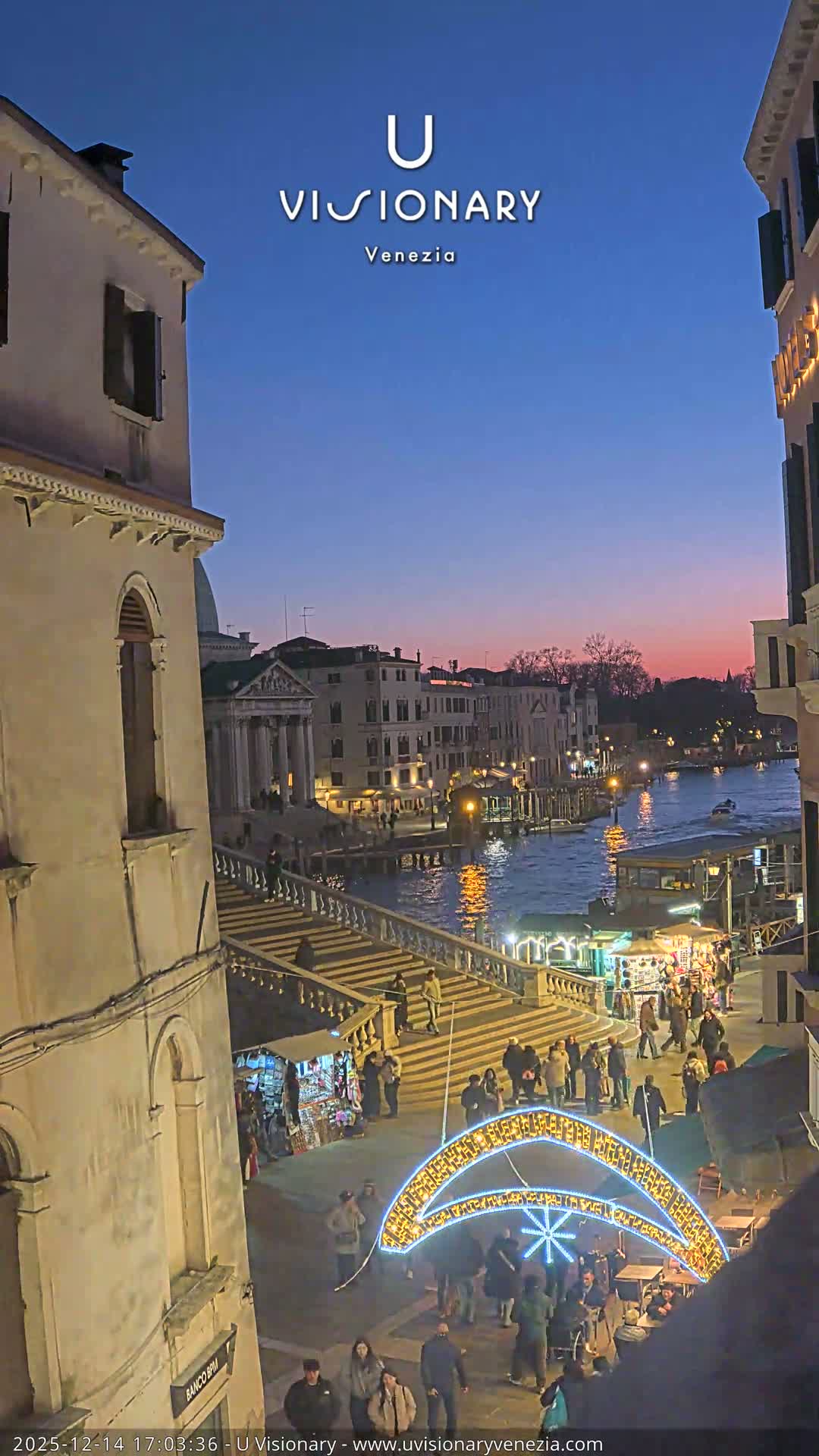 On a clear and sunny morning, a bustling scene in Venice captures people ascending and descending a grand stone bridge over a canal filled with boats, while market stalls and pedestrians animate the paved area below.