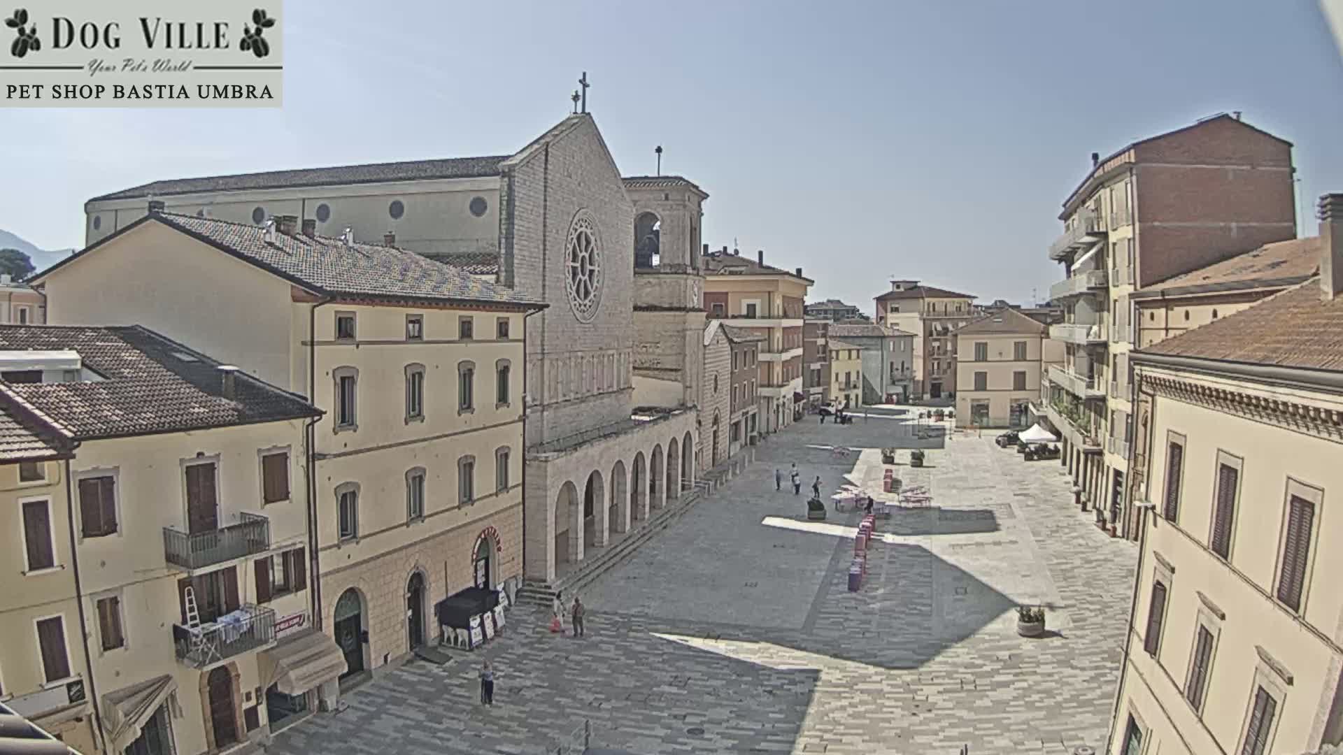 A large town square, under sunny skies, is surrounded by multi-story buildings, one of which is a church.
