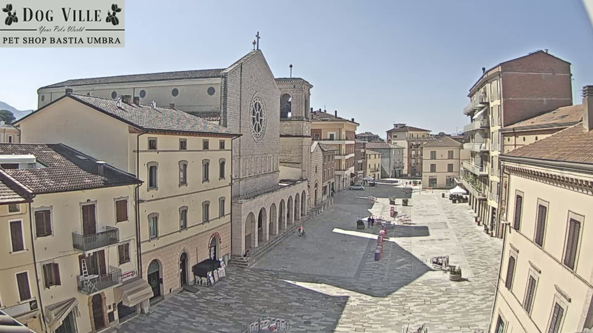 A large town square on a sunny day is surrounded by buildings of varying heights and architectural styles, with a prominent church at the center.