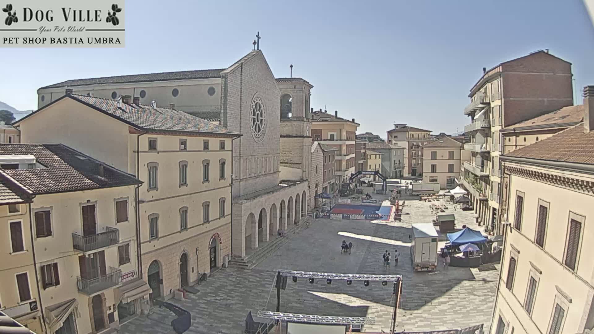 A large town square, under sunny skies, is filled with people and event preparations near a prominent church.