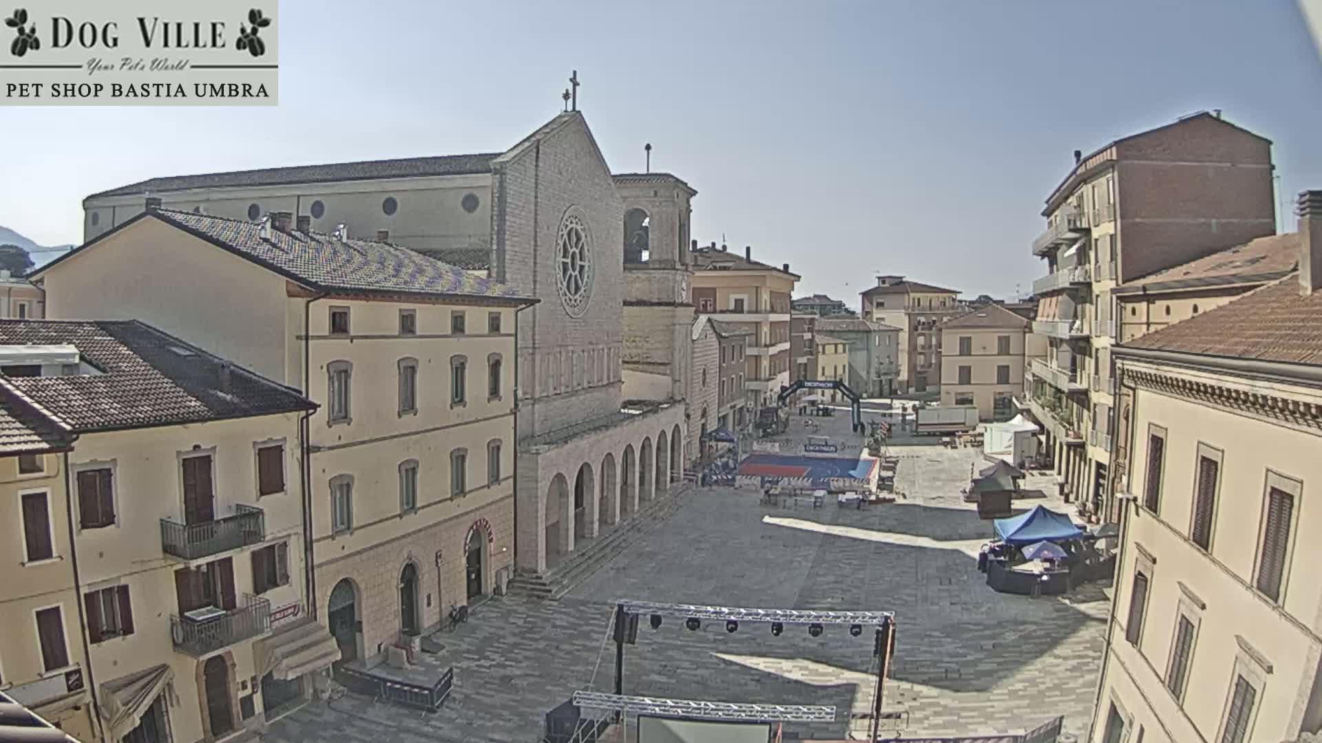 A sunny town square is shown with a large stone church in the background, several buildings surrounding the square, and a stage and various tents set up in the center.
