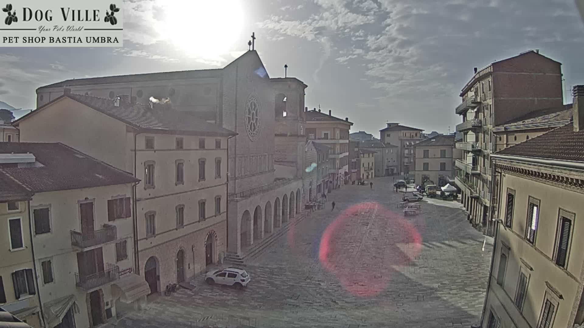 A high vantage point shows a quiet European town square with a prominent stone church featuring a rose window and an arched colonnade, surrounded by multi-story buildings, a few pedestrians, and a parked car, all under a partly cloudy sky with bright sunshine.