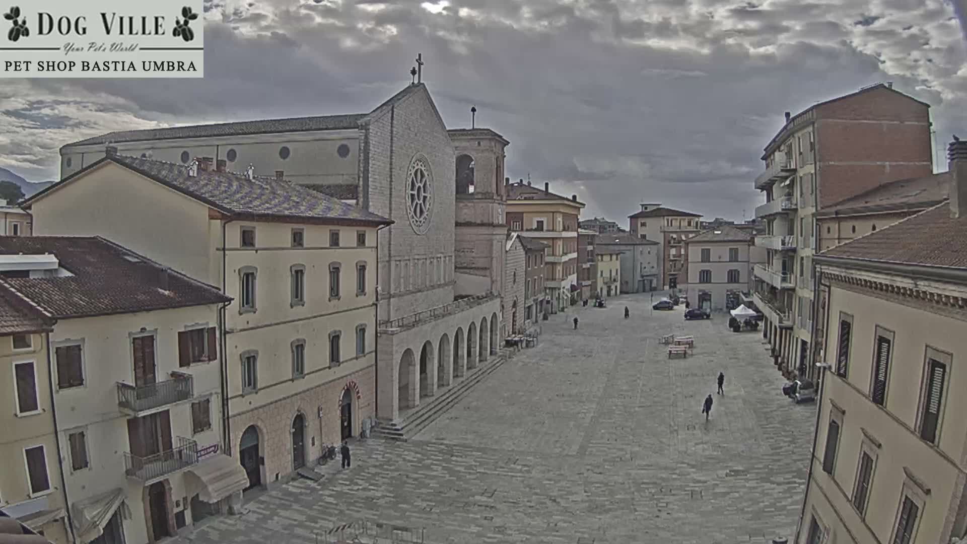 A high vantage point shows a quiet European town square with a prominent stone church featuring a rose window and an arched colonnade, surrounded by multi-story buildings, a few pedestrians, and a parked car, all under a partly cloudy sky with bright sunshine.