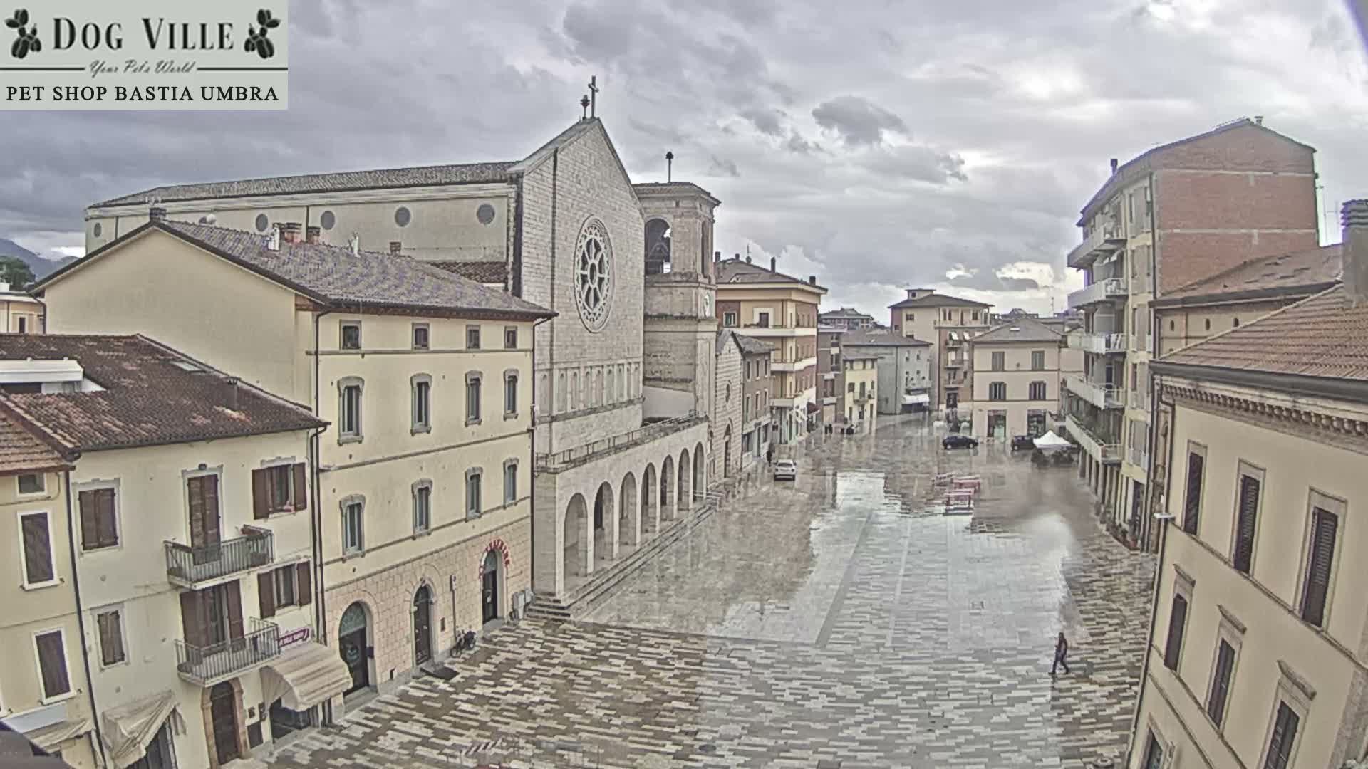 A historic European town square, featuring a prominent stone church and surrounding buildings, appears wet and reflective from recent rain under a heavily overcast sky.
