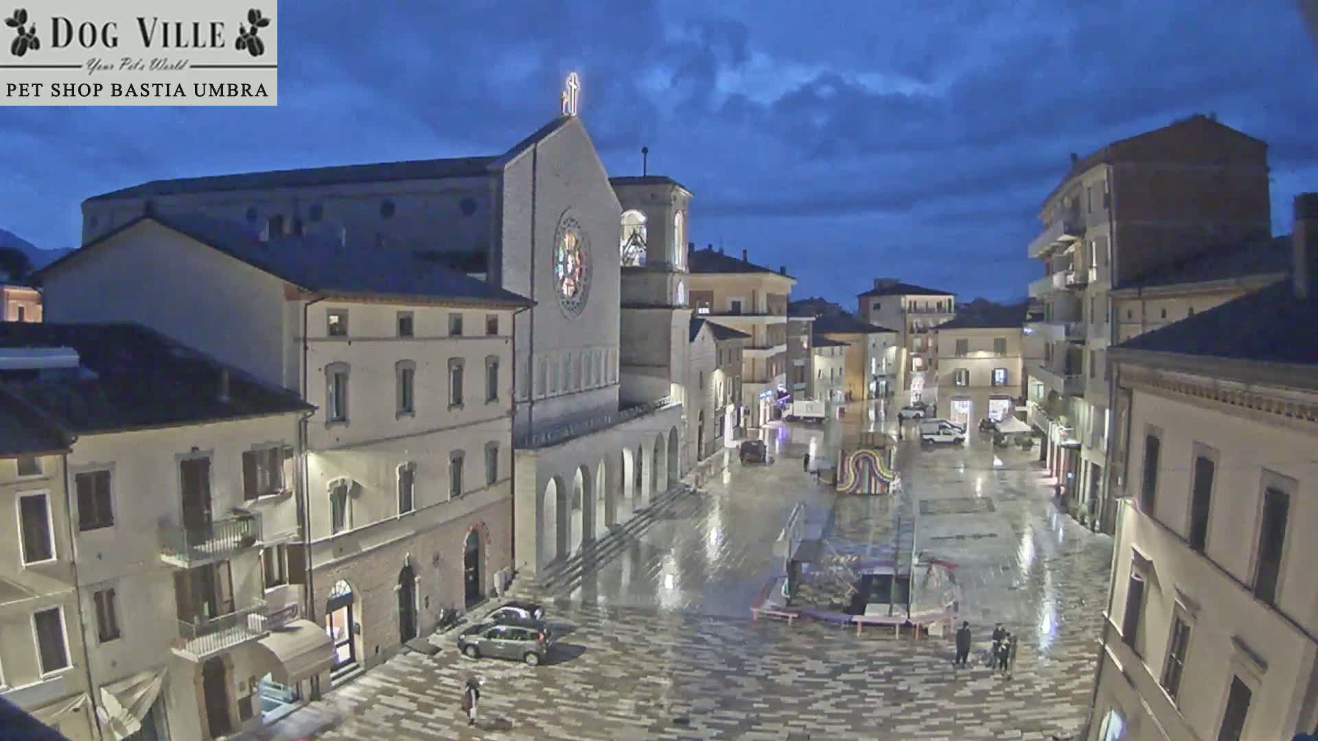 A wet city square with a prominent illuminated church and surrounding buildings reflects streetlights under a dark, cloudy sky at dusk, with a few people and vehicles present on the damp pavement after rain.