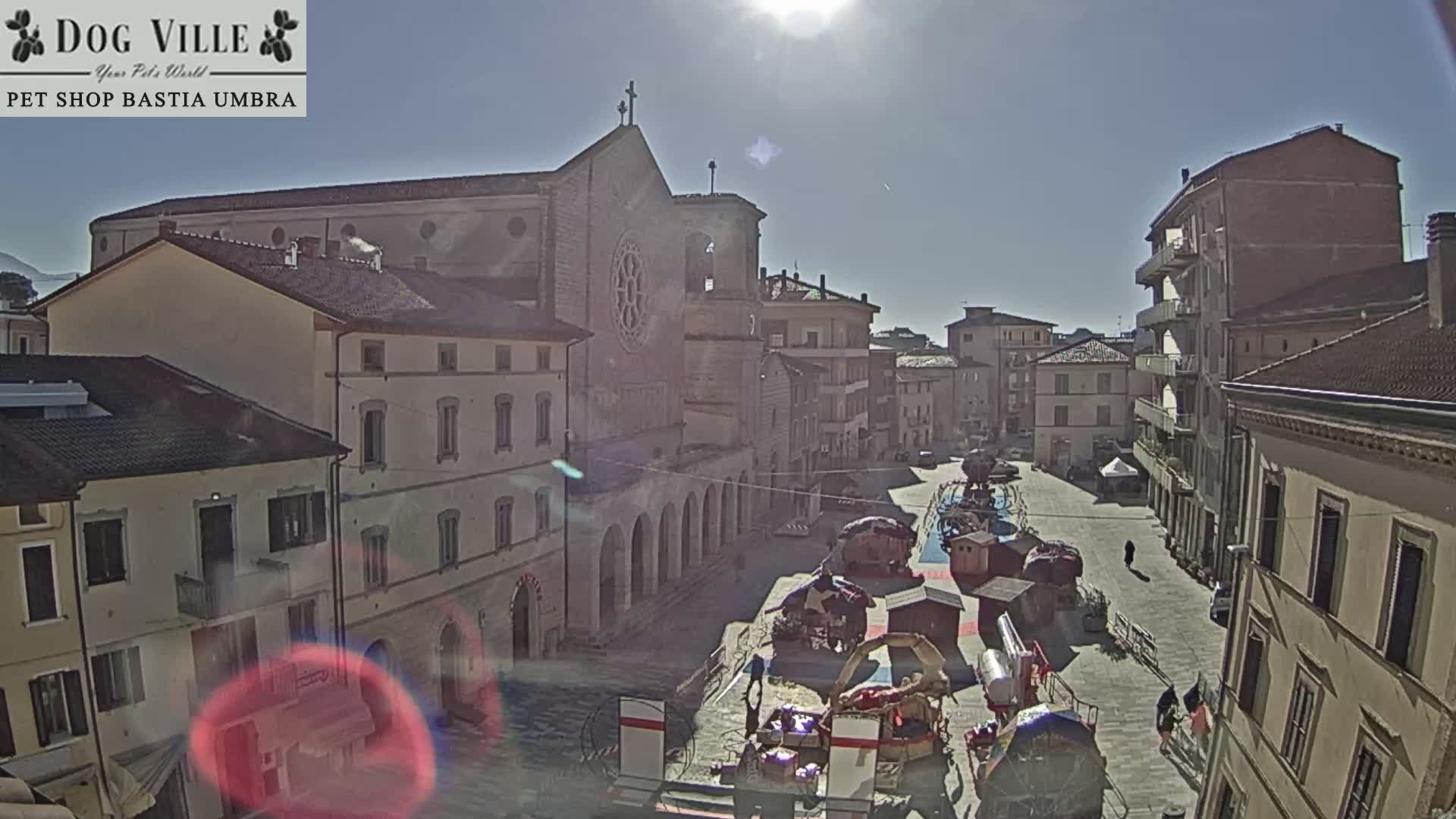 Under a bright, clear, sunny sky, a historic European town square features a large church with a prominent rose window and bell tower, surrounded by multi-story buildings, and a collection of temporary market stalls with a few pedestrians.