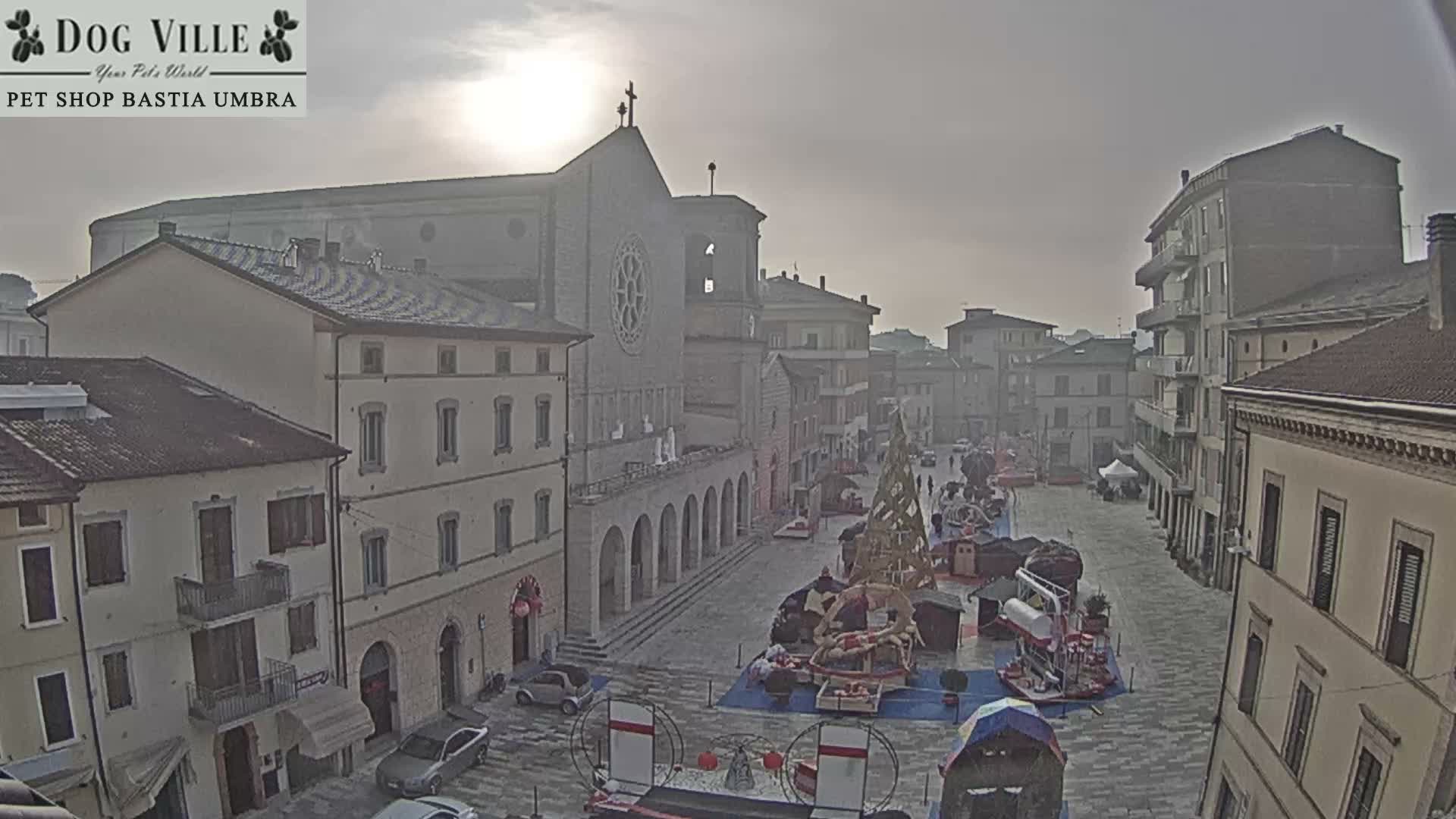 An overcast day hangs over a European town square featuring a prominent cathedral, surrounding buildings, and a festive market area decorated with stalls and a large wooden tree-like structure.