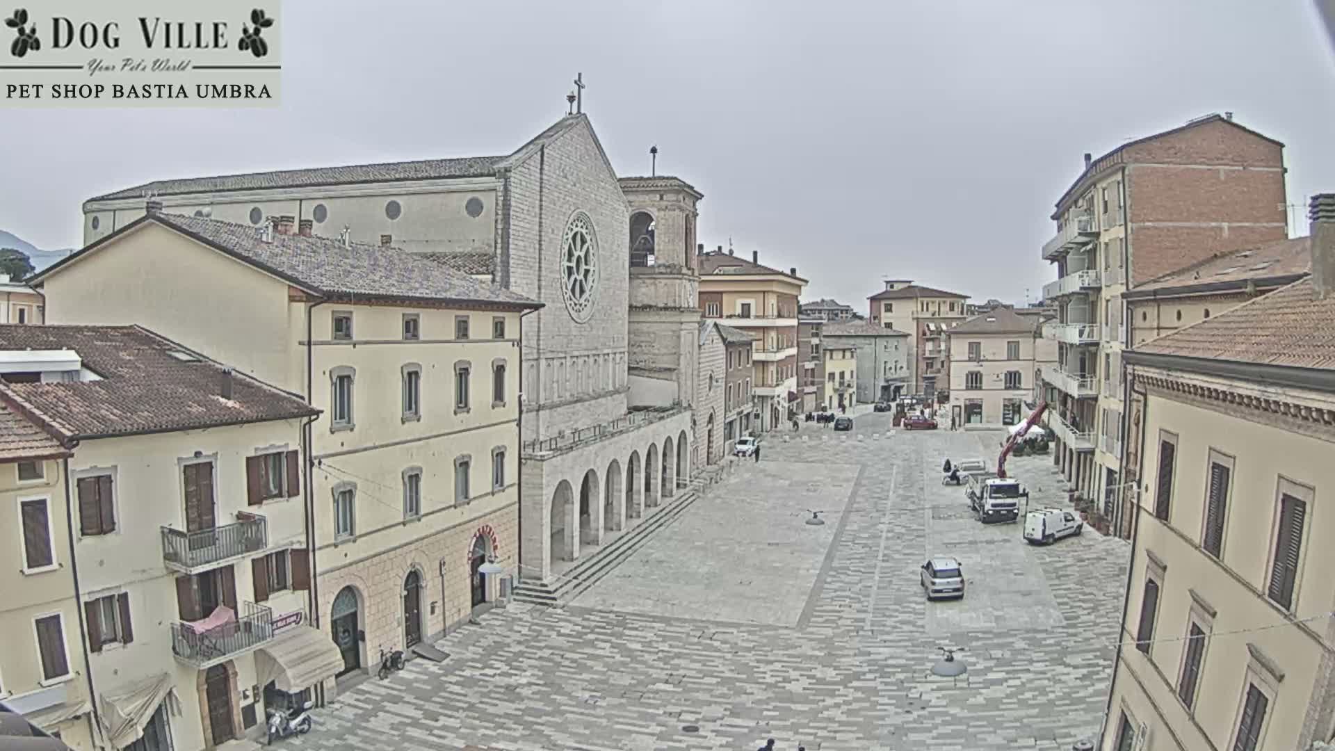 An overcast day hangs over a European town square featuring a prominent cathedral, surrounding buildings, and a festive market area decorated with stalls and a large wooden tree-like structure.