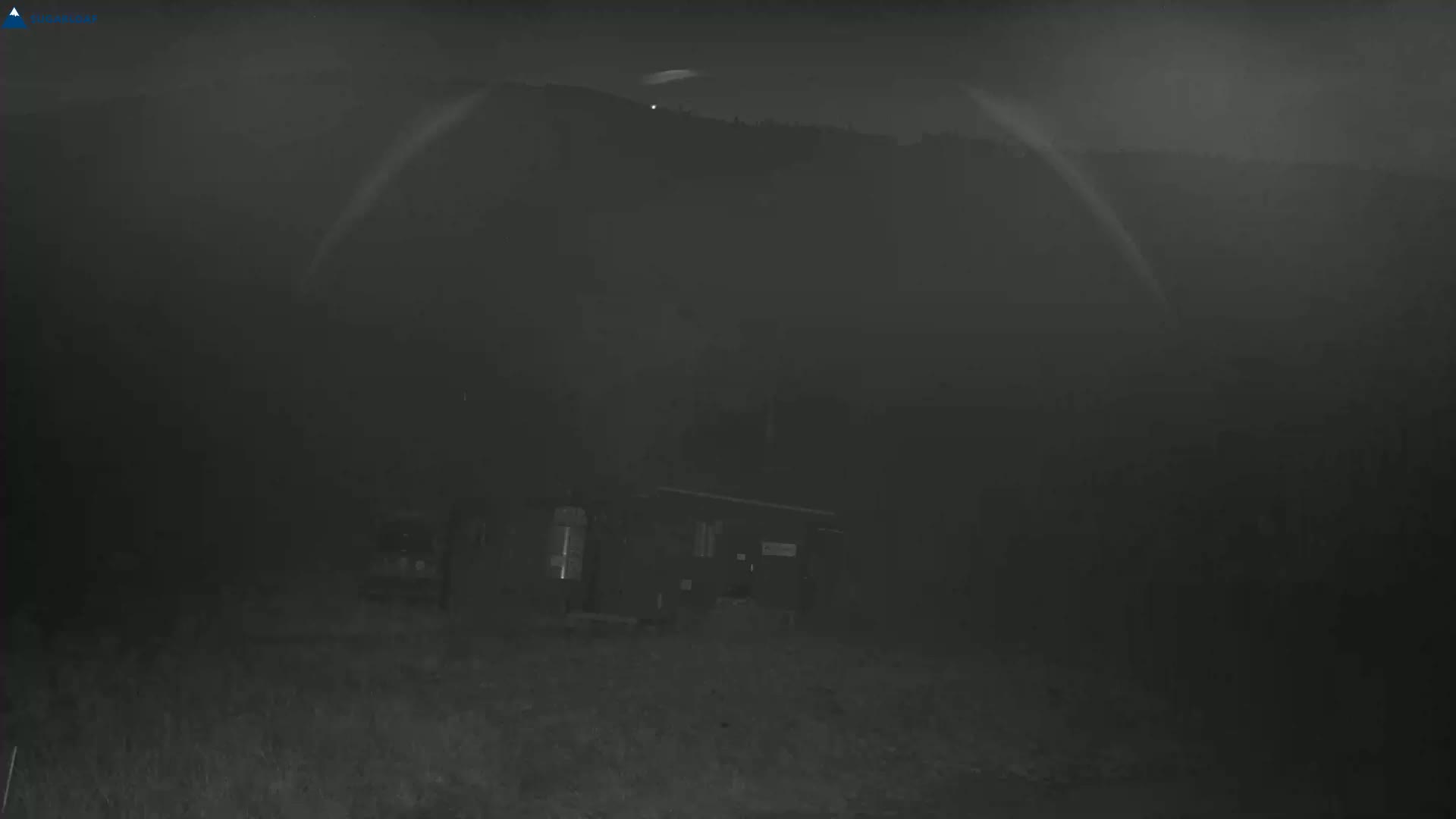 A small building sits in a foggy, dark night scene, with hills visible in the background under a mostly clear sky.
