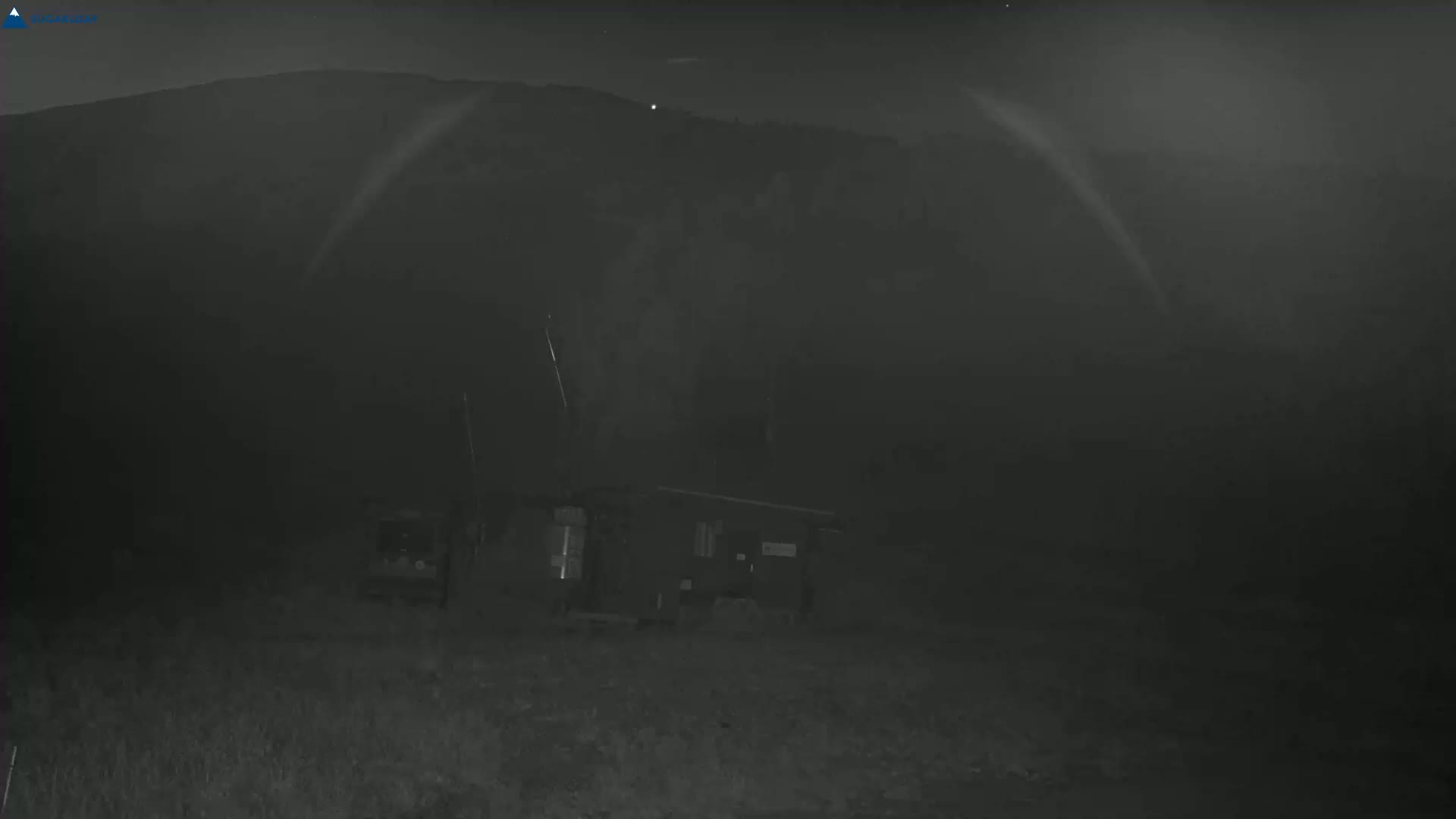 A small structure sits in a foggy, dark, nighttime landscape before a distant mountain range under a mostly clear sky.
