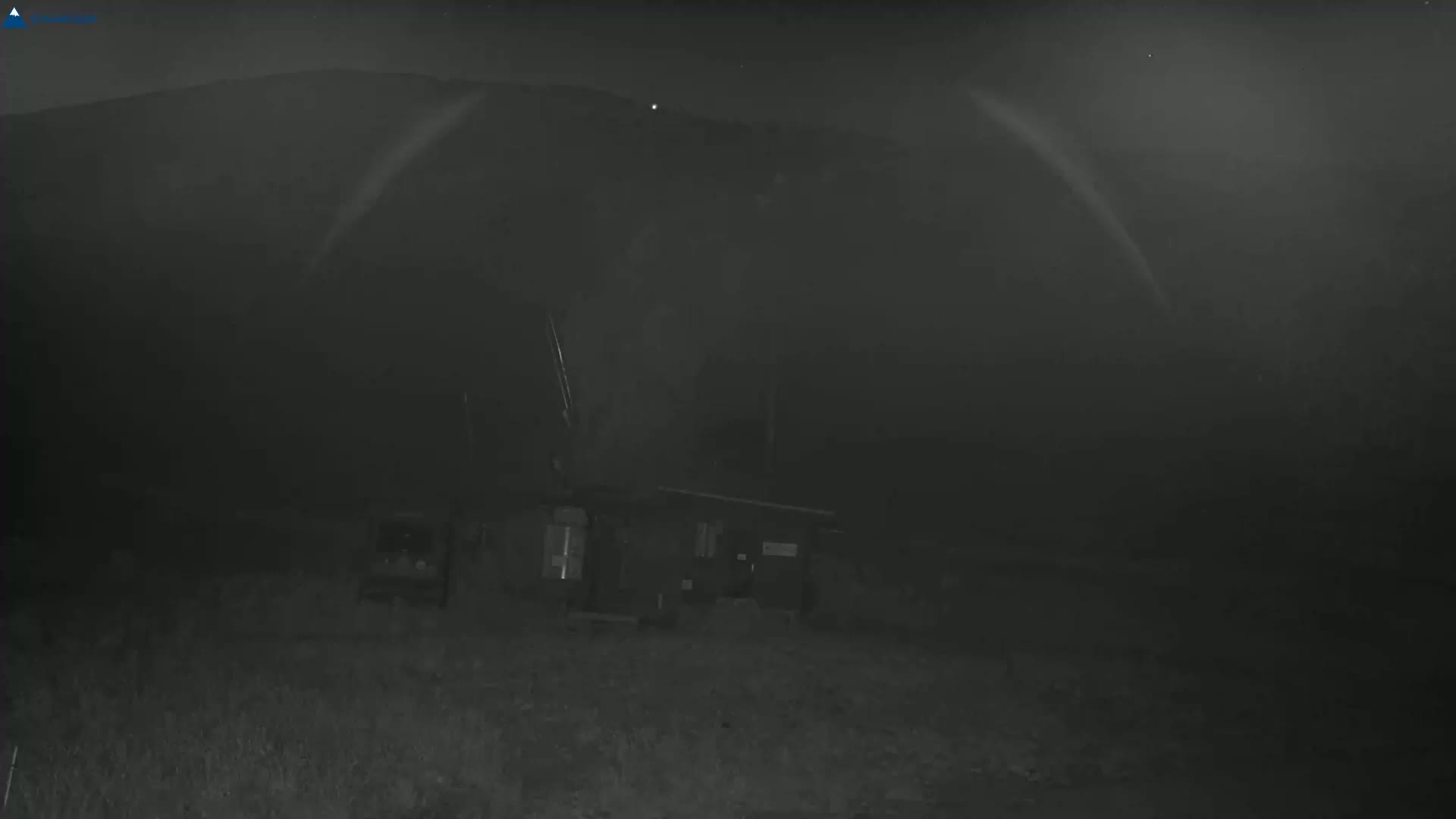 A small building is visible in the foreground under a dark, possibly foggy or hazy, night sky with a mountain in the background.