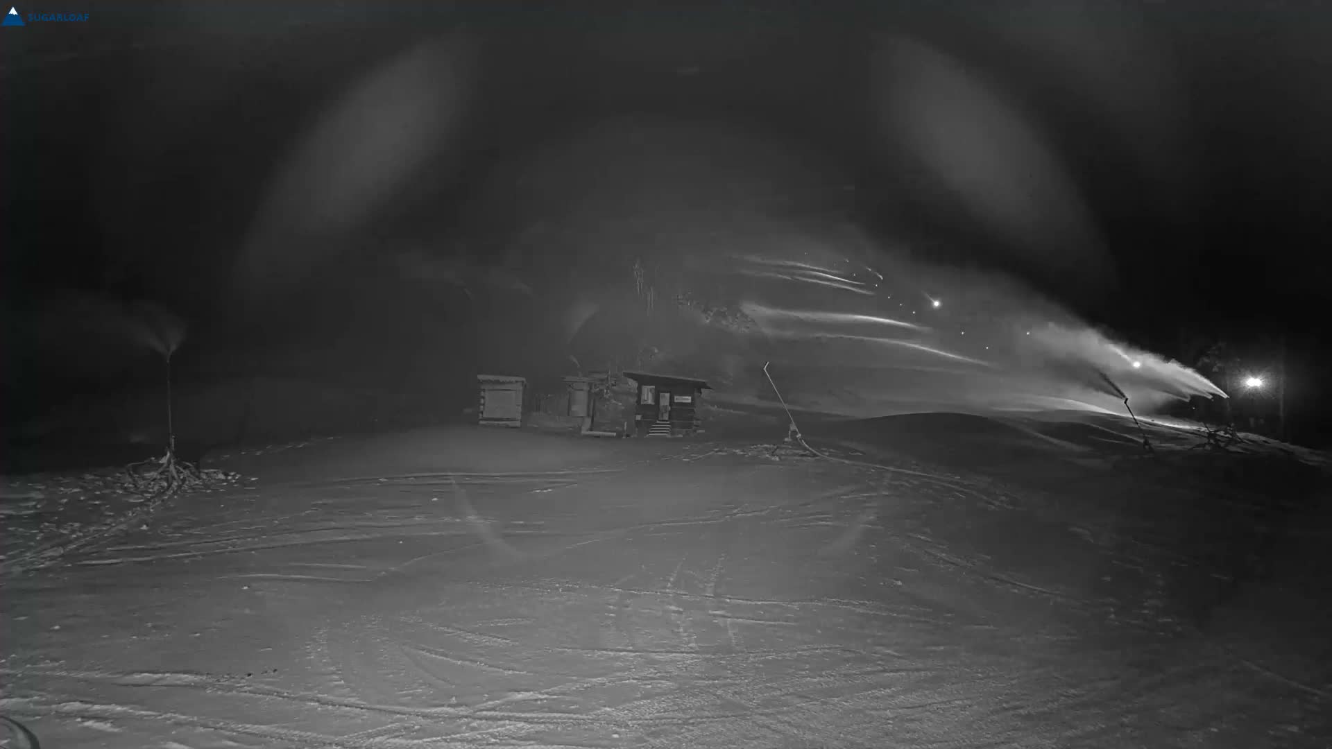 A snow-covered ski slope is visible at night, illuminated by lights, with multiple snowmaking machines actively blowing snow under clear, cold conditions.