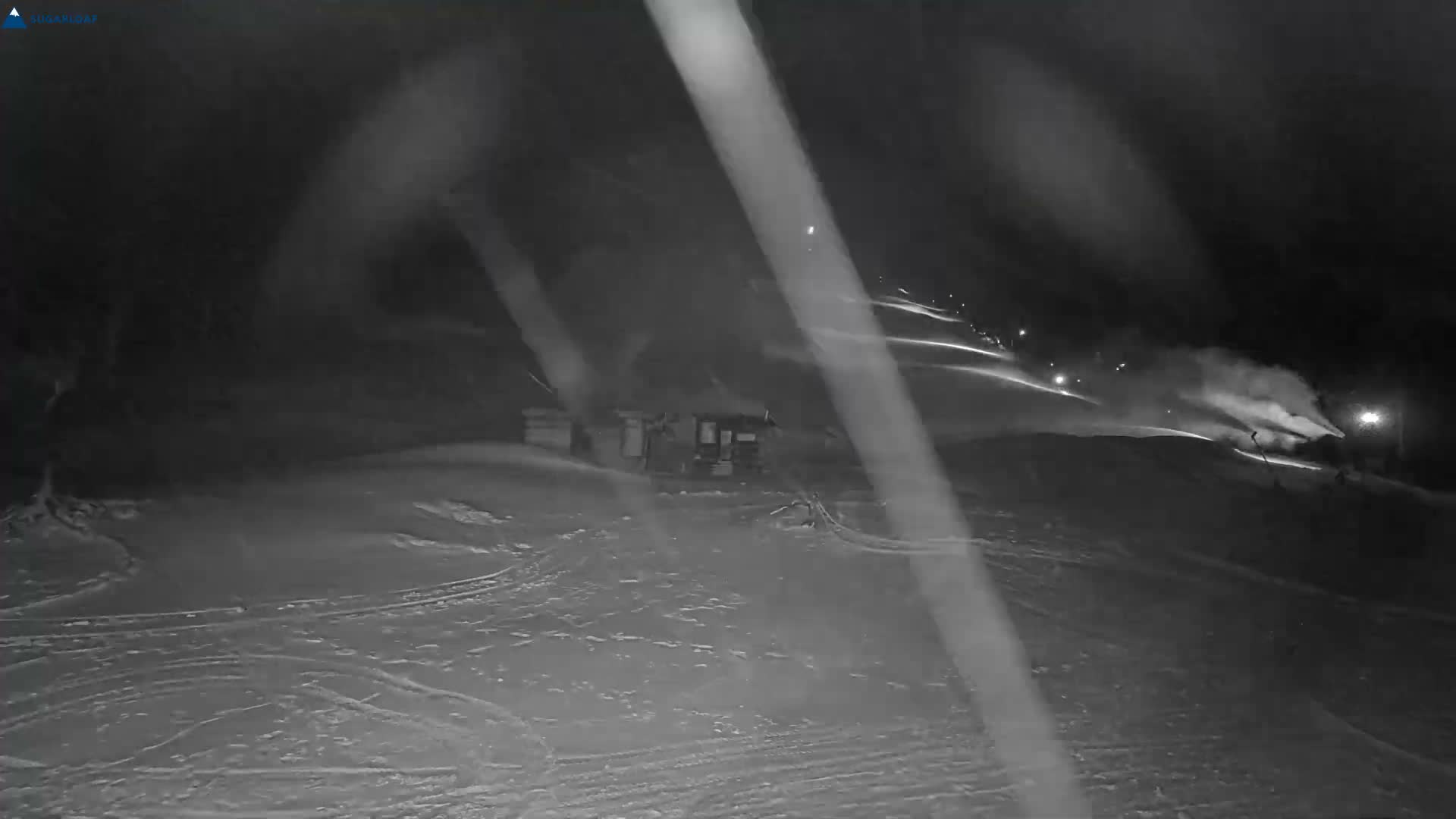 A cold, clear night at a snow-covered ski resort shows an active snow cannon blowing snow on the right, with illuminated ski slopes extending into the distance and small buildings in the middle ground.