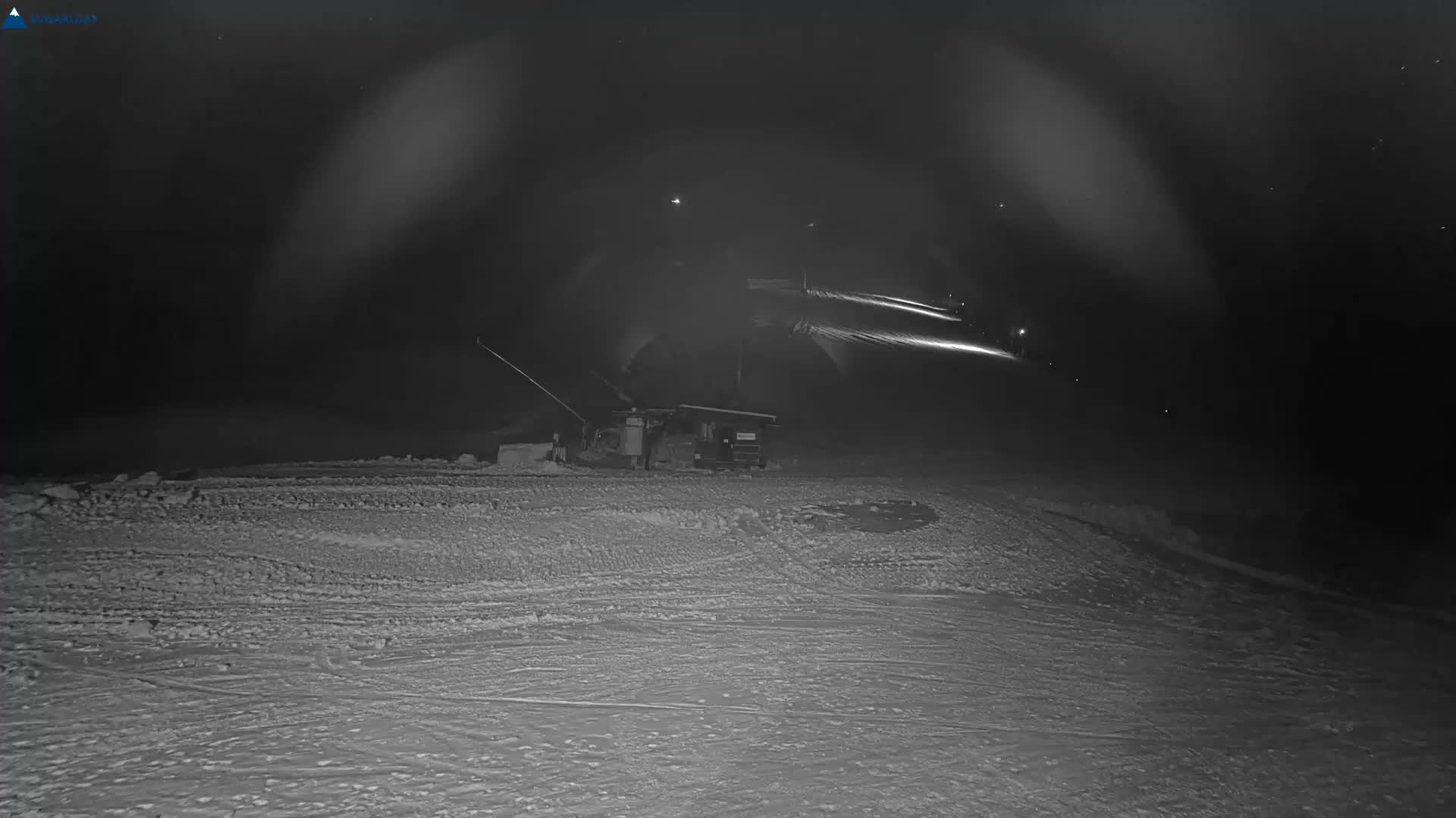 A snowy ski slope is brightly illuminated by artificial lights at night, revealing groomed snow, active snowmaking equipment, and a clear, cold sky.