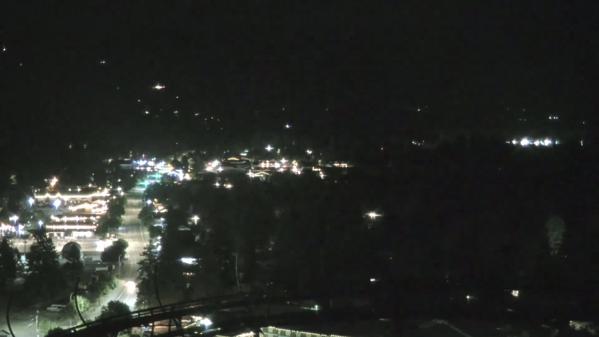 A nighttime, high-angle view of a town illuminated by scattered lights, with a roller coaster visible in the lower portion of the frame.