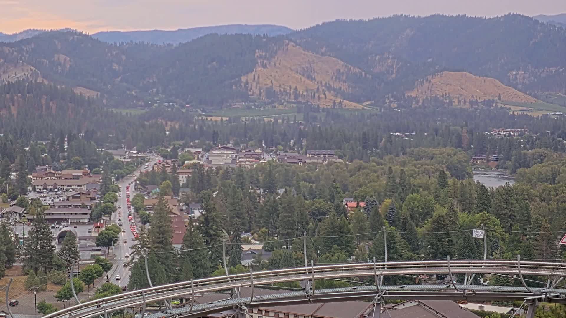 A mountain town nestled in a valley, with a river visible in the distance, under hazy skies.
