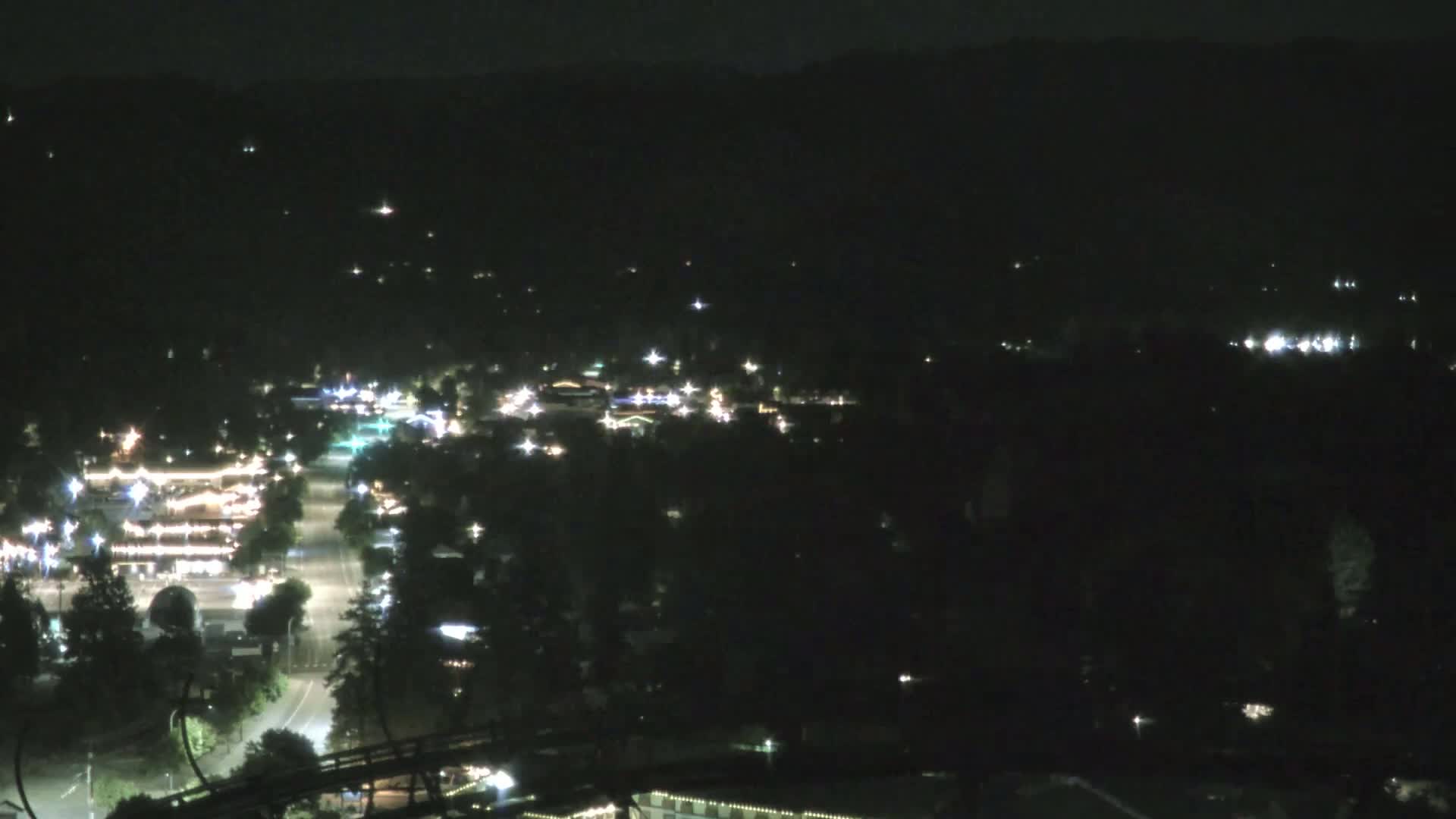 A nighttime, high-angle view shows a town with scattered lights against a dark, tree-covered landscape.