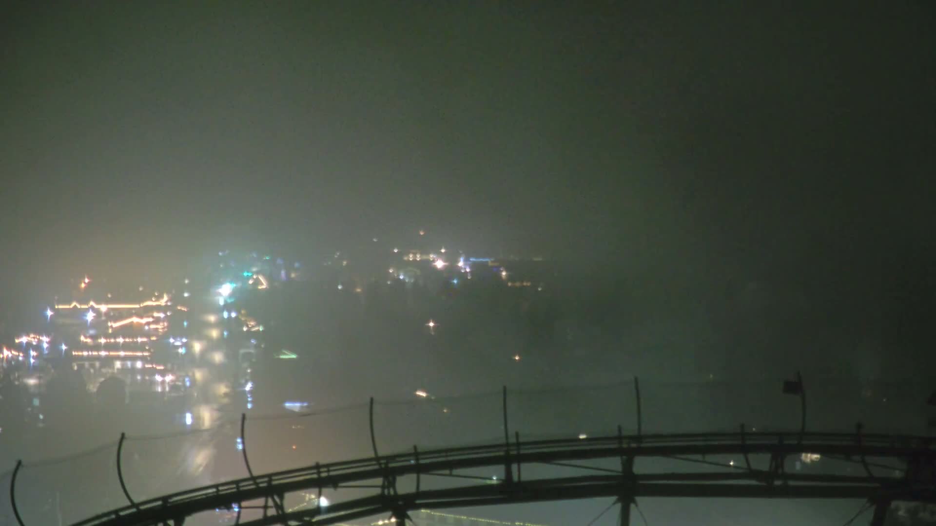 A hazy and foggy night obscures the distant landscape, revealing only the diffused, colorful lights of a sprawling resort or town and a dark, curved roller coaster track in the foreground.