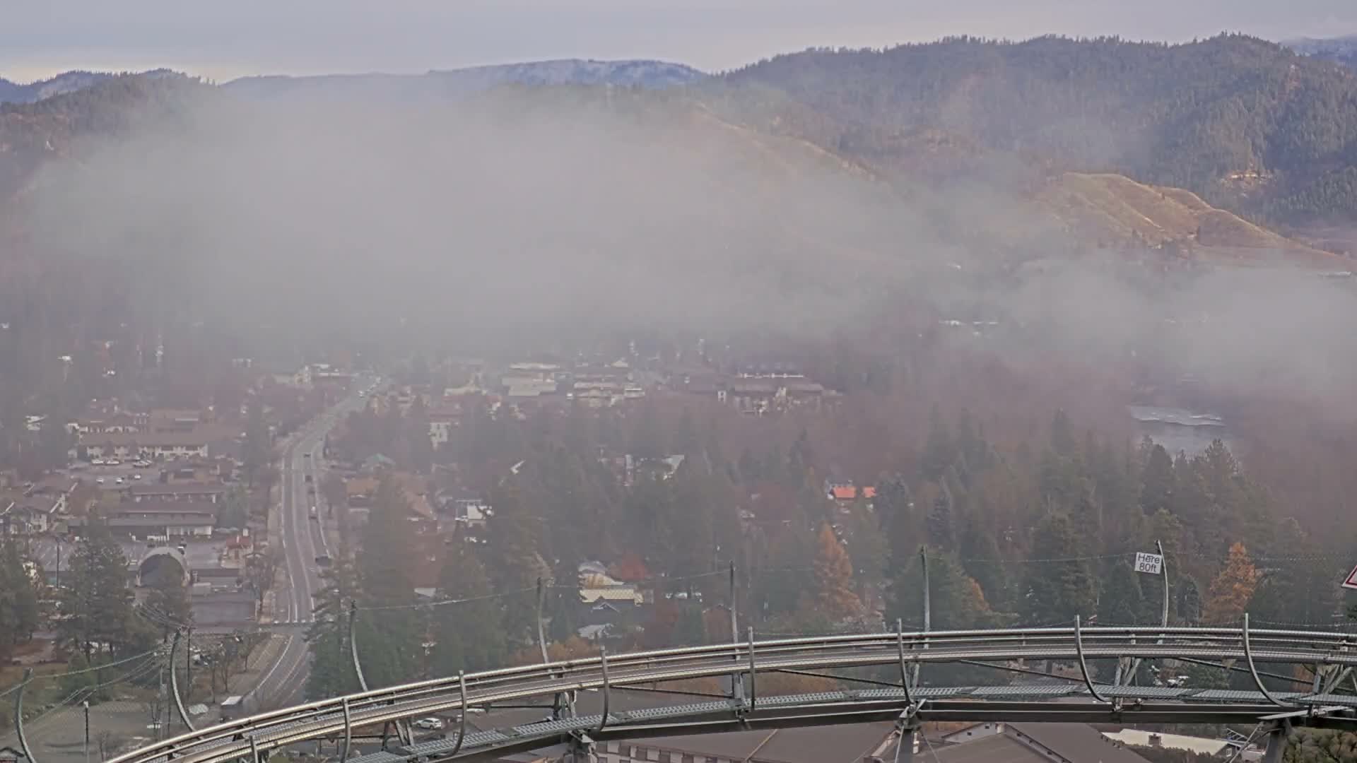 A hazy and foggy night obscures the distant landscape, revealing only the diffused, colorful lights of a sprawling resort or town and a dark, curved roller coaster track in the foreground.