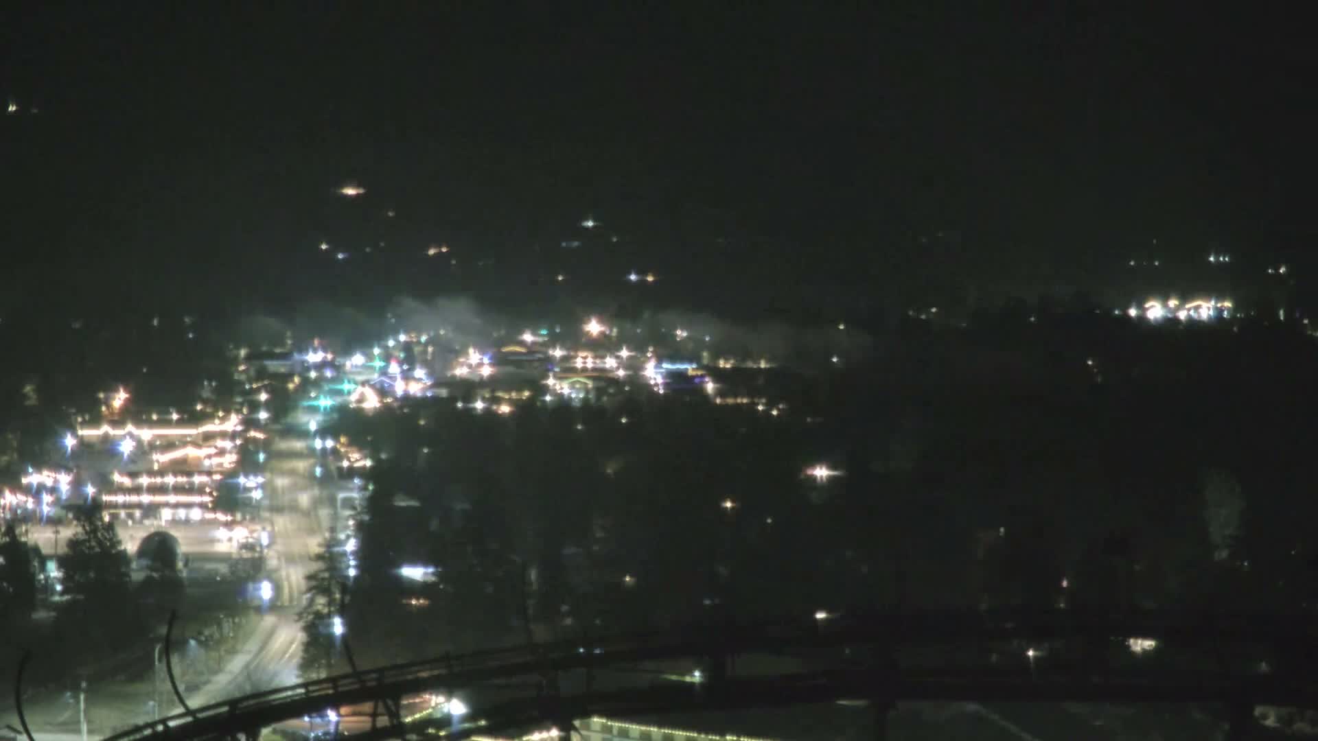 A panoramic night view showcases a brightly lit town or resort area, adorned with numerous lights and some visible atmospheric haze, from an elevated perspective with a dark track structure in the foreground.