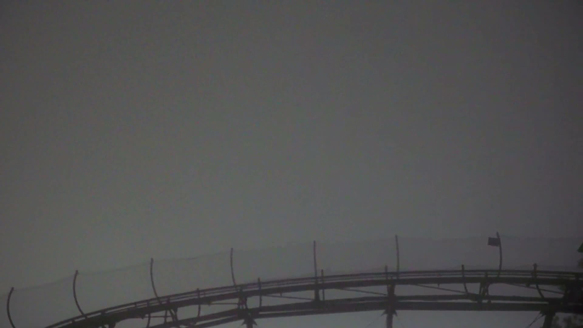 A dark, elevated wooden roller coaster track with safety netting is silhouetted against a uniformly grey and hazy sky under overcast conditions.