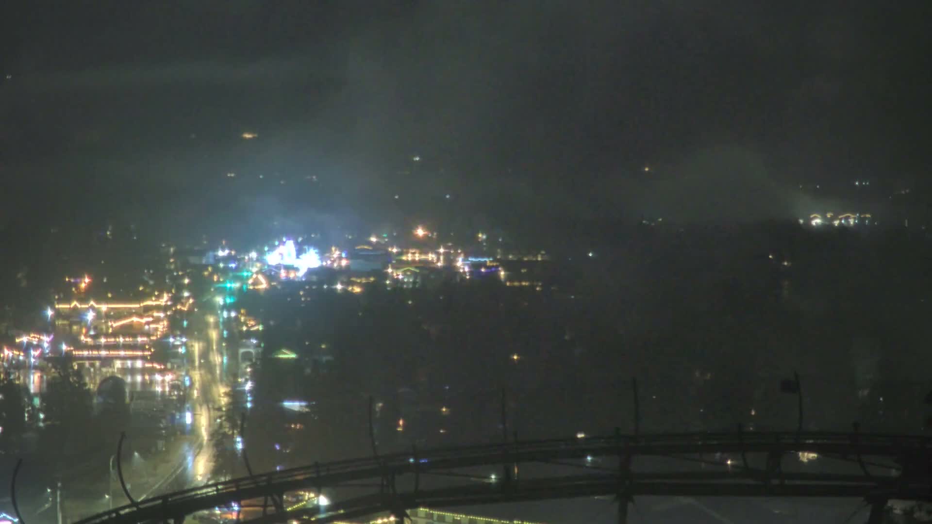 A foggy night scene from above shows a brightly illuminated town with wet streets reflecting lights, framed by a dark foreground structure resembling a roller coaster track.