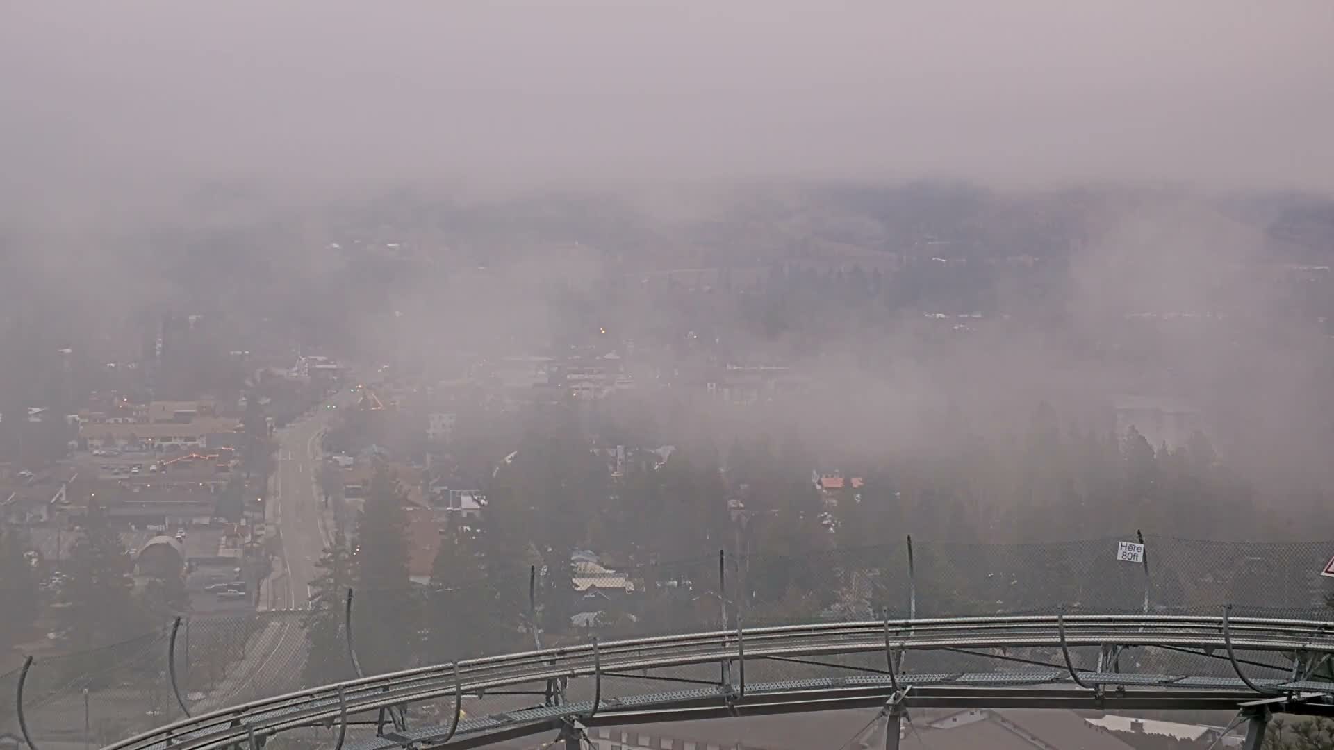 A foggy night scene from above shows a brightly illuminated town with wet streets reflecting lights, framed by a dark foreground structure resembling a roller coaster track.