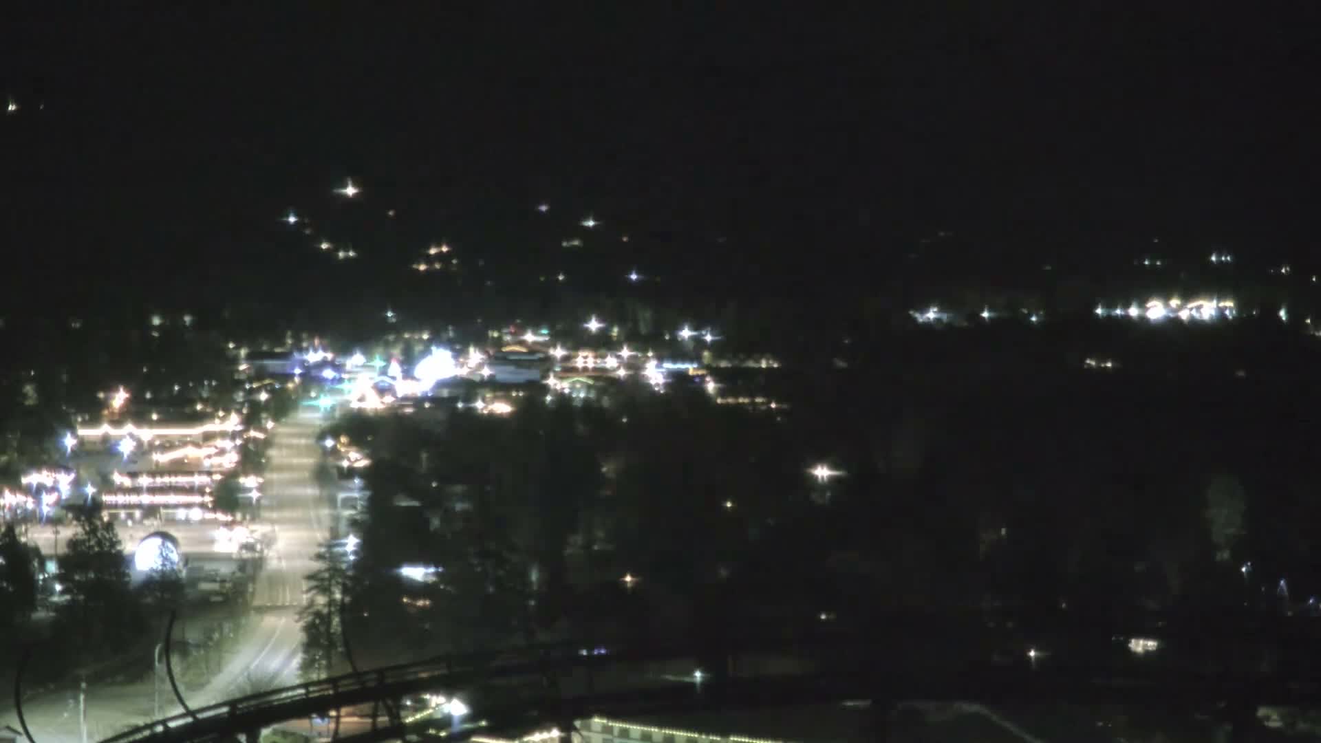 A foggy night scene from above shows a brightly illuminated town with wet streets reflecting lights, framed by a dark foreground structure resembling a roller coaster track.