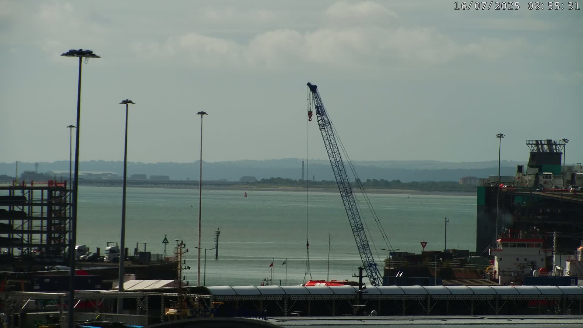 A large crane stands over a calm body of water, next to a dock with various vehicles and structures, under a mostly cloudy sky.