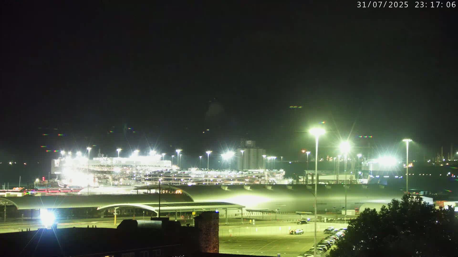 A brightly lit port at night, with a large vehicle carrier ship docked, under a dark, clear sky.