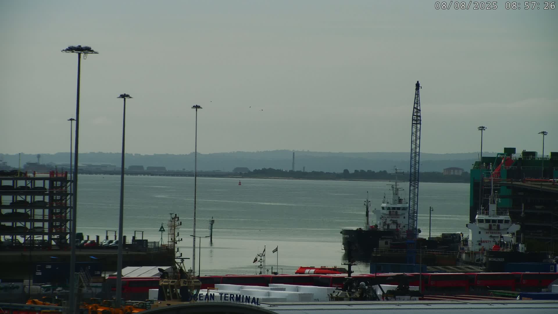 A hazy, overcast day reveals a port with several ships docked, a multi-story parking structure, and distant hills across a calm body of water.