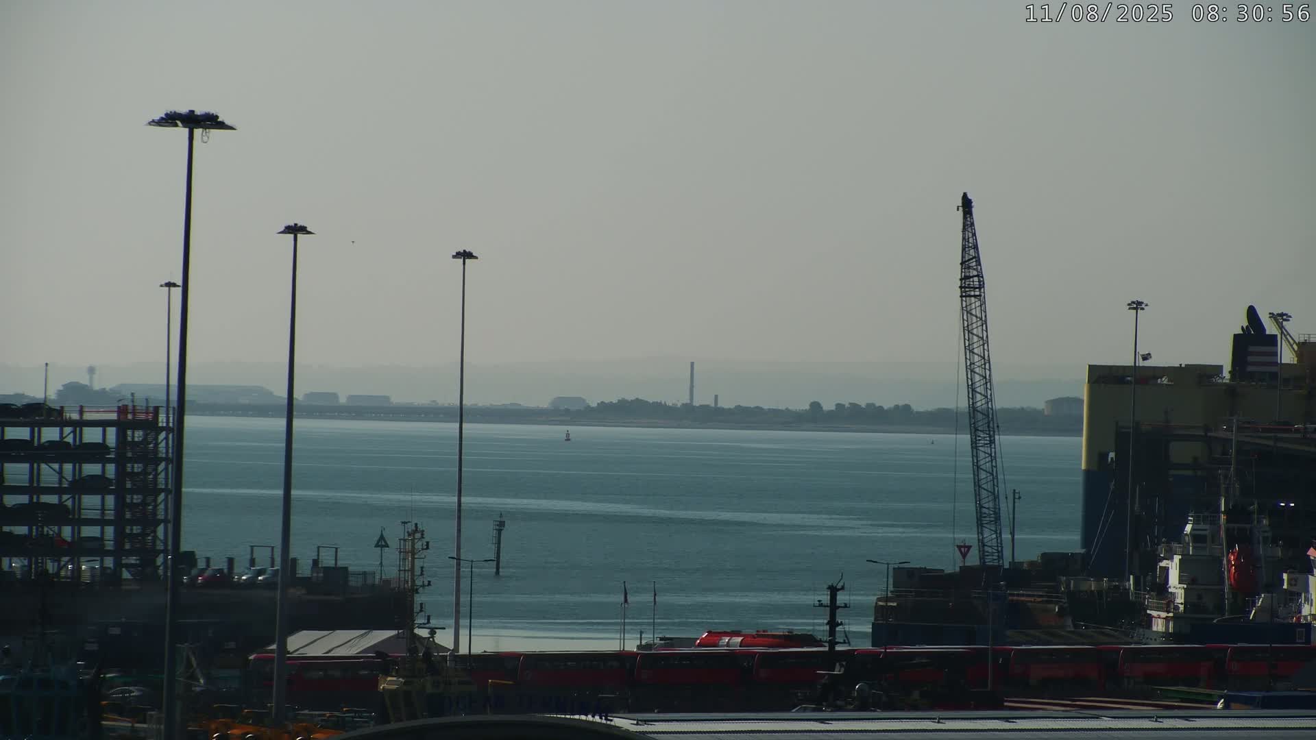 A hazy, light-colored body of water stretches to a distant shoreline, featuring a port with cranes, vehicles, and tall light posts in the foreground under a muted sky.